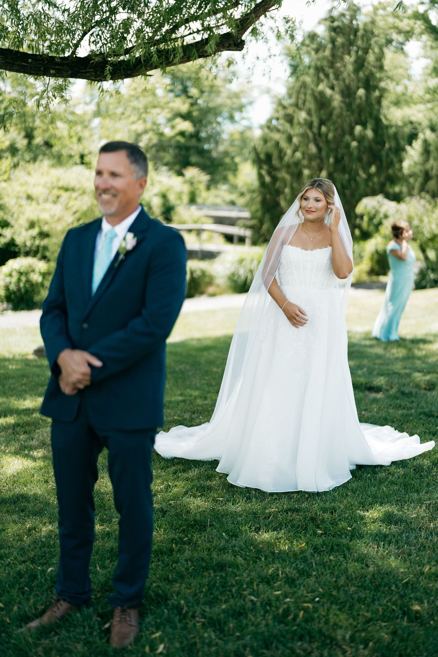 Bride in white gown and veil approaches smiling groom in navy suit during outdoor first look ceremony.