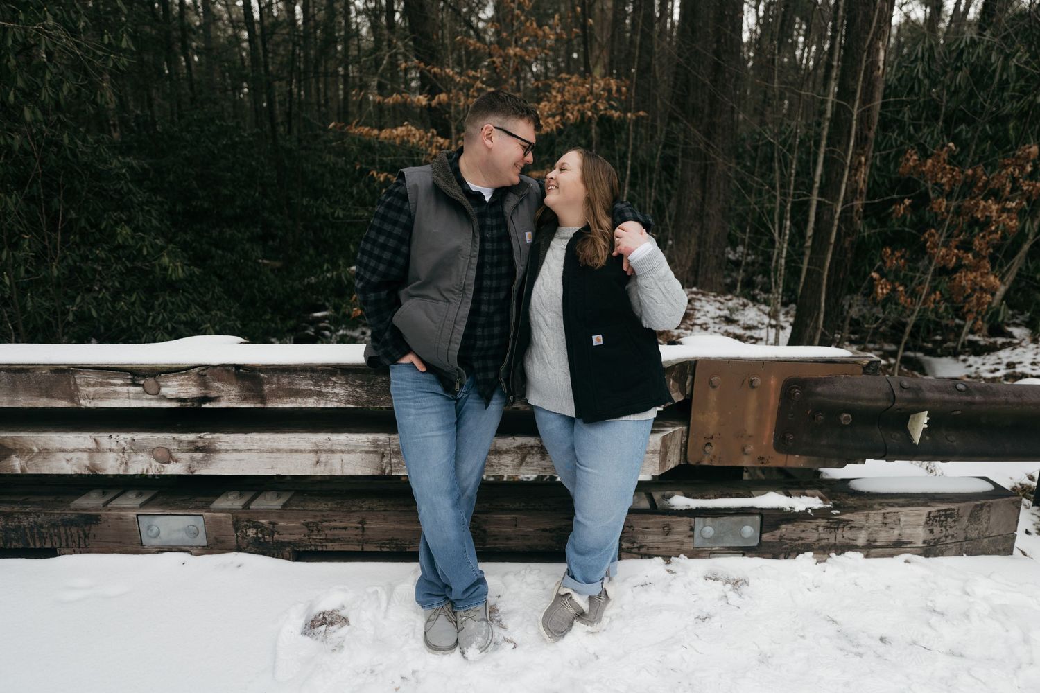 A couple shares a sweet moment while sitting on a snow-covered wooden railing in the woods.