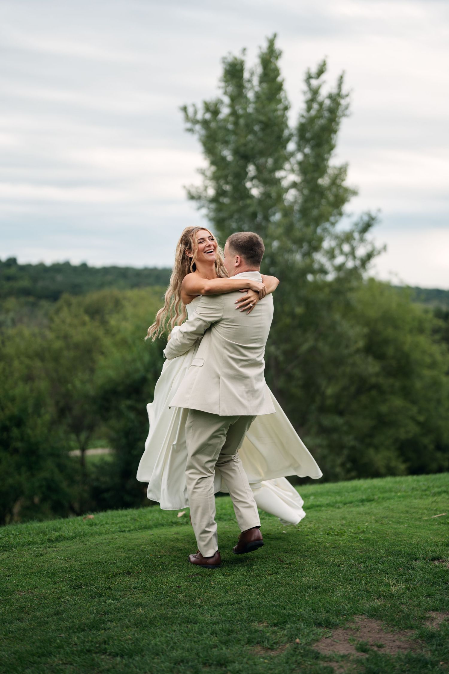 Joyful bride in white dress being lifted by groom in beige suit on green hillside with trees in background.