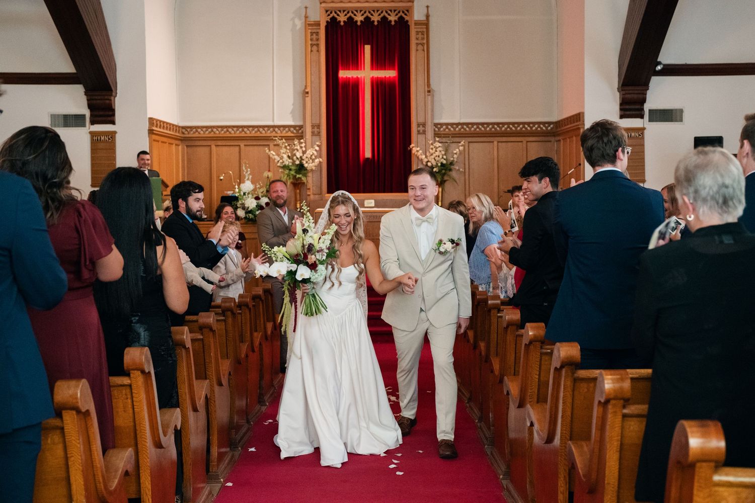 Bride and groom walk down red carpet aisle of historic church after wedding ceremony, guests cheering on both sides.