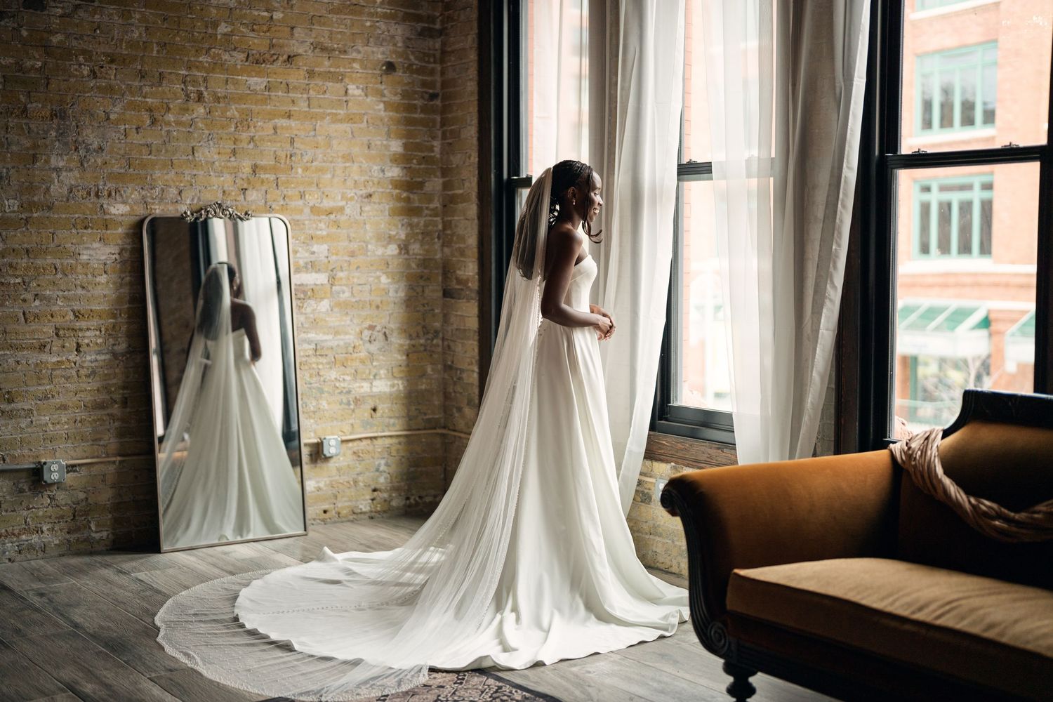 Bride in elegant white wedding gown with long veil stands by tall window, brick wall and vintage sofa in background.