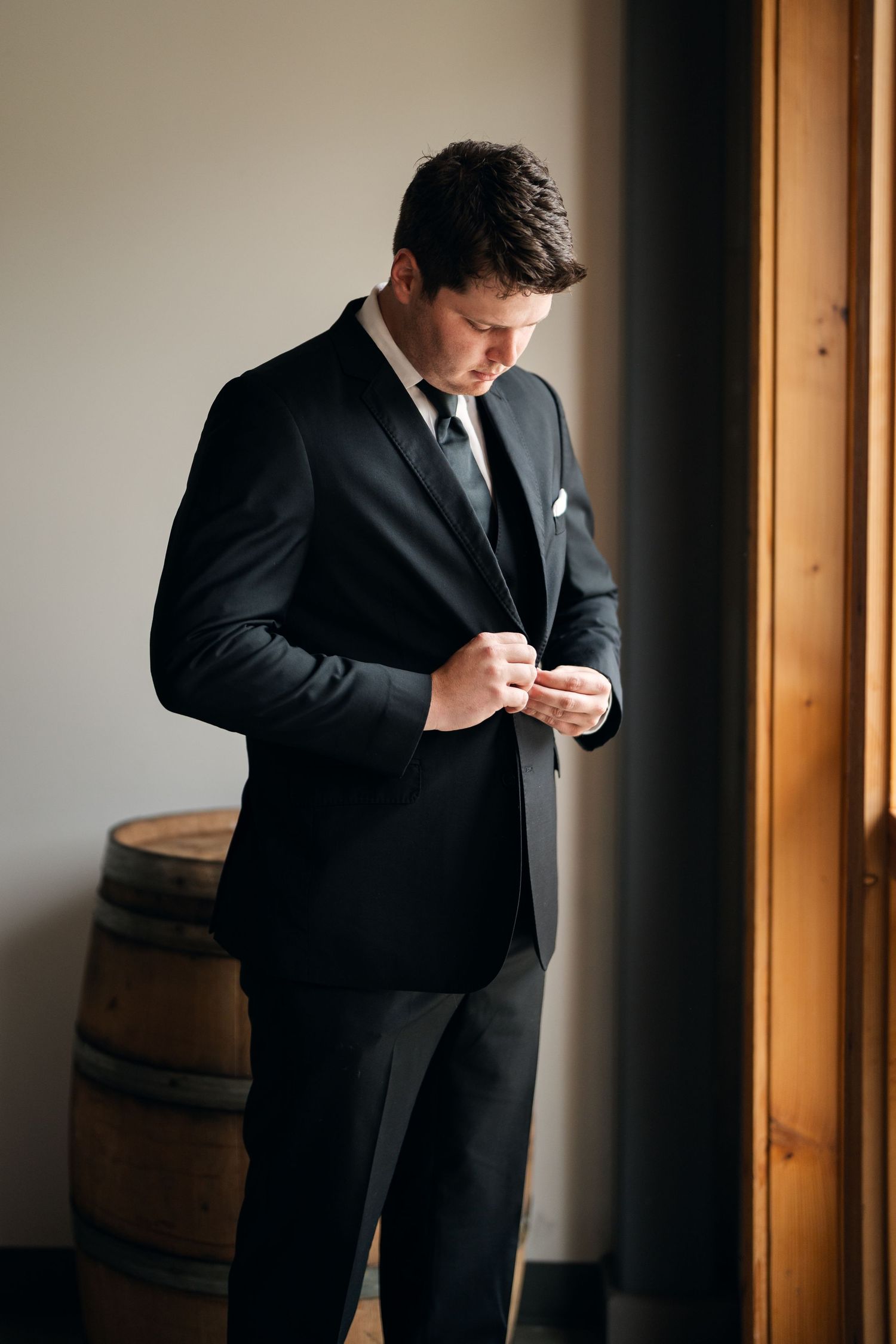 Groom in black suit adjusting cufflinks by window with wooden barrel backdrop.