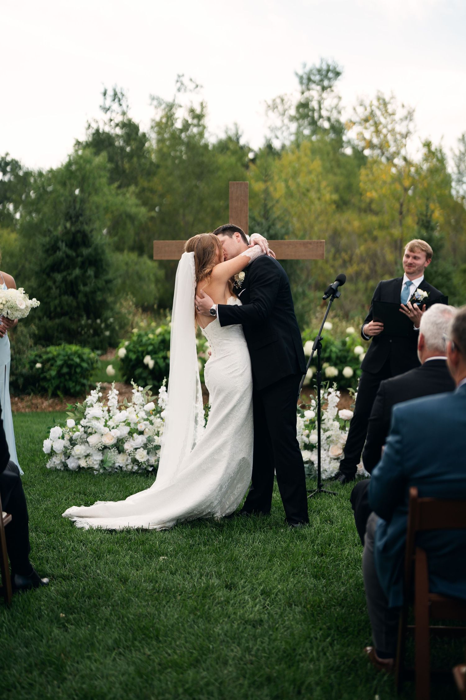 Bride and groom share first kiss at outdoor wedding ceremony with wooden cross and white floral arrangements.