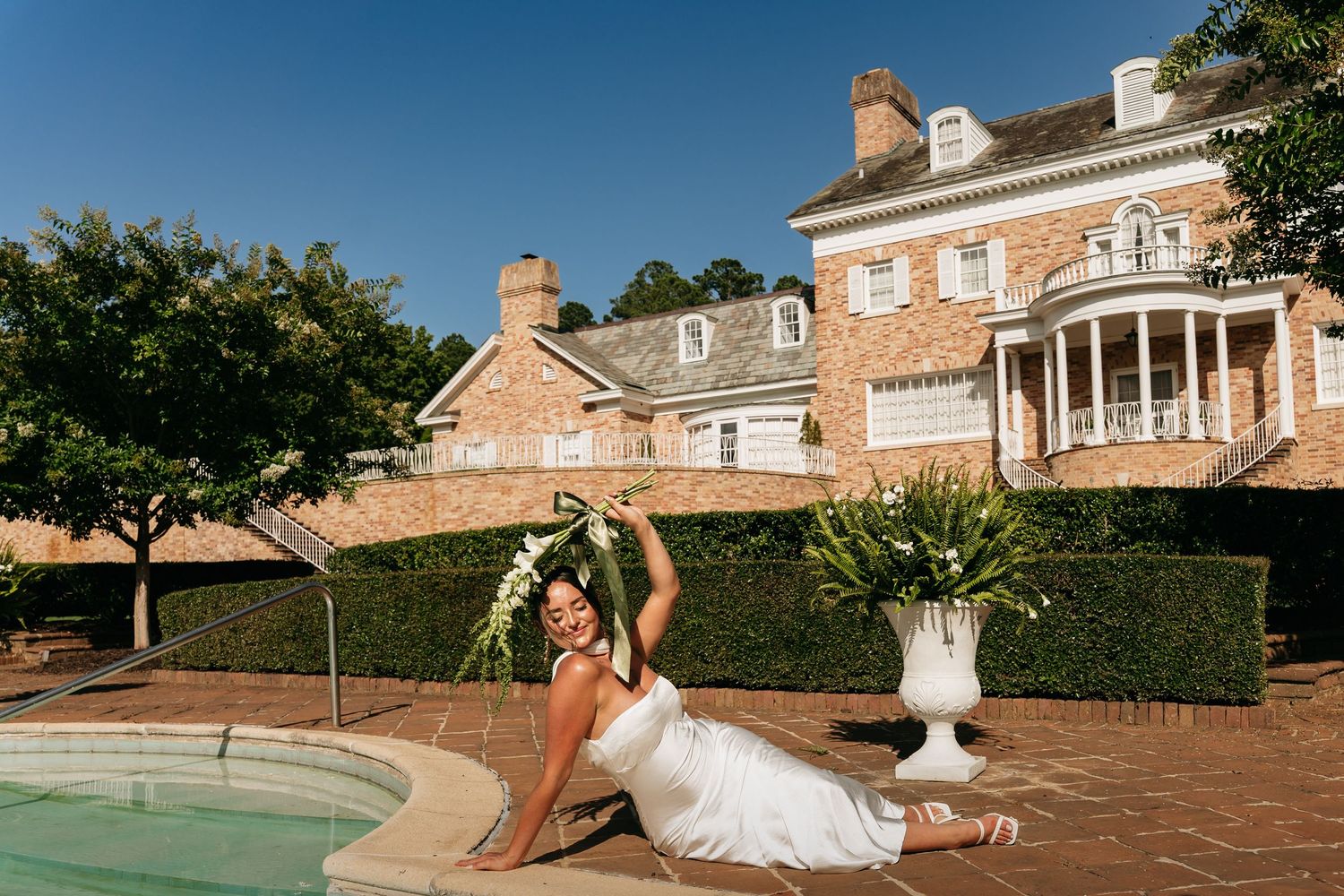 Bride in white gown poses gracefully by a pool at a luxurious brick estate mansion on a sunny day.