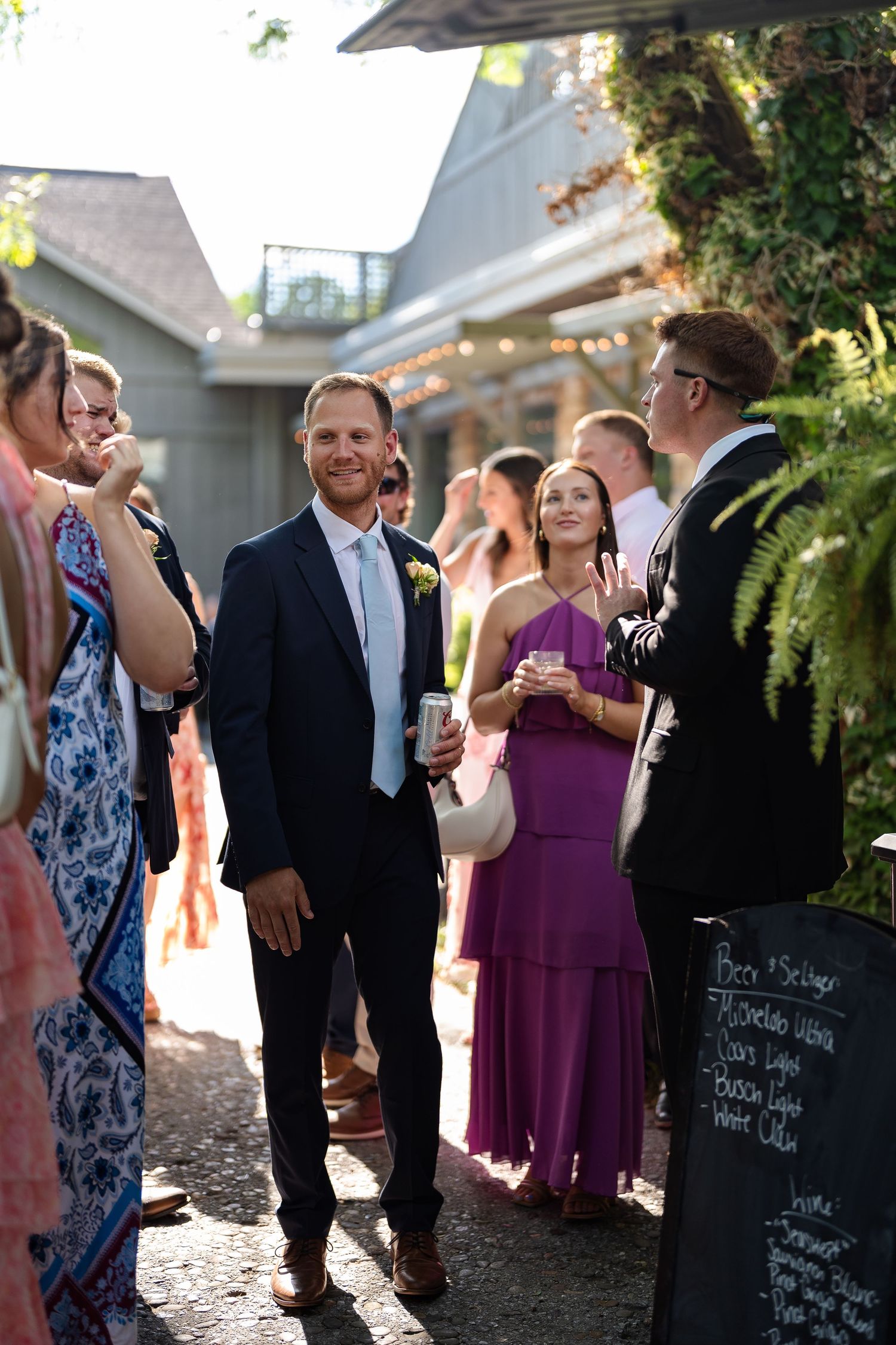 Wedding guests mingling outdoors at rustic venue with string lights and lush greenery.