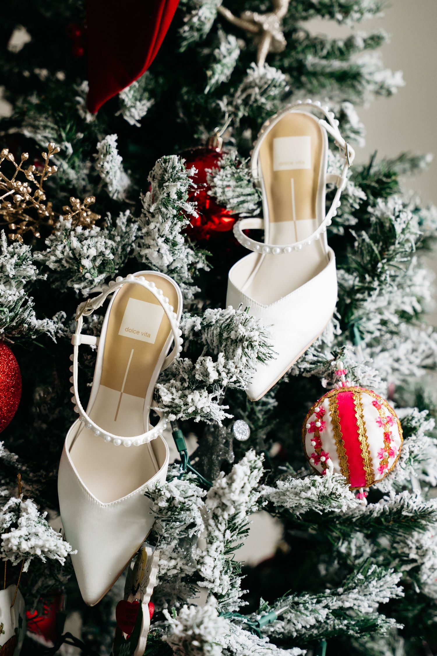 White high heel shoes hanging as Christmas tree ornaments on a snowy evergreen tree with red decorations.