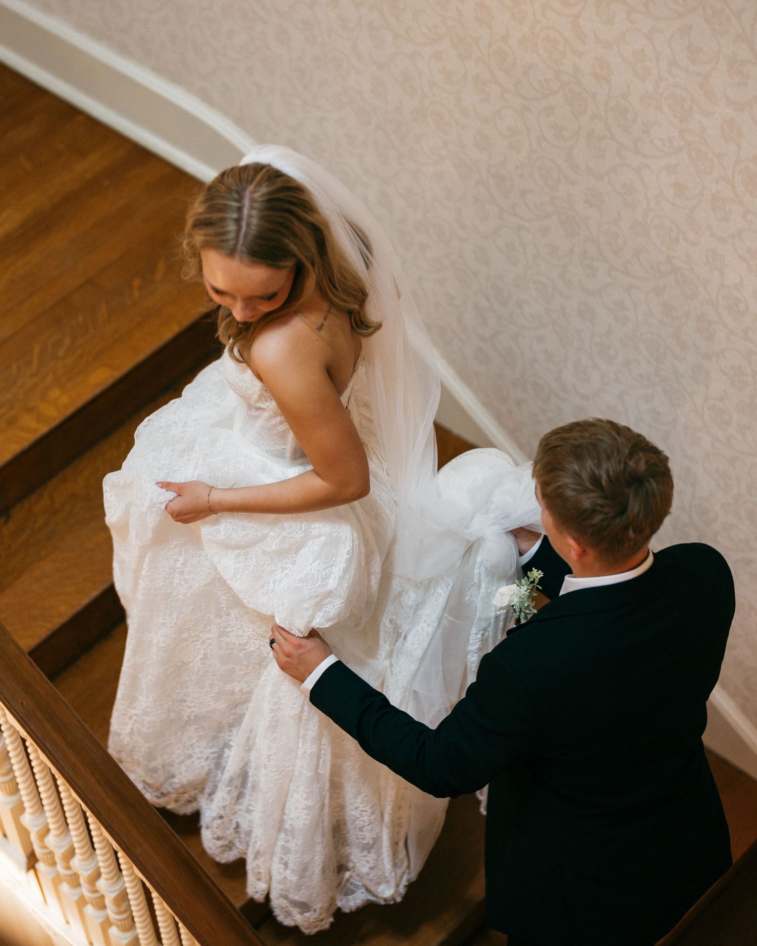 Aerial view of groom in navy suit helping bride in lace wedding dress on wooden staircase.