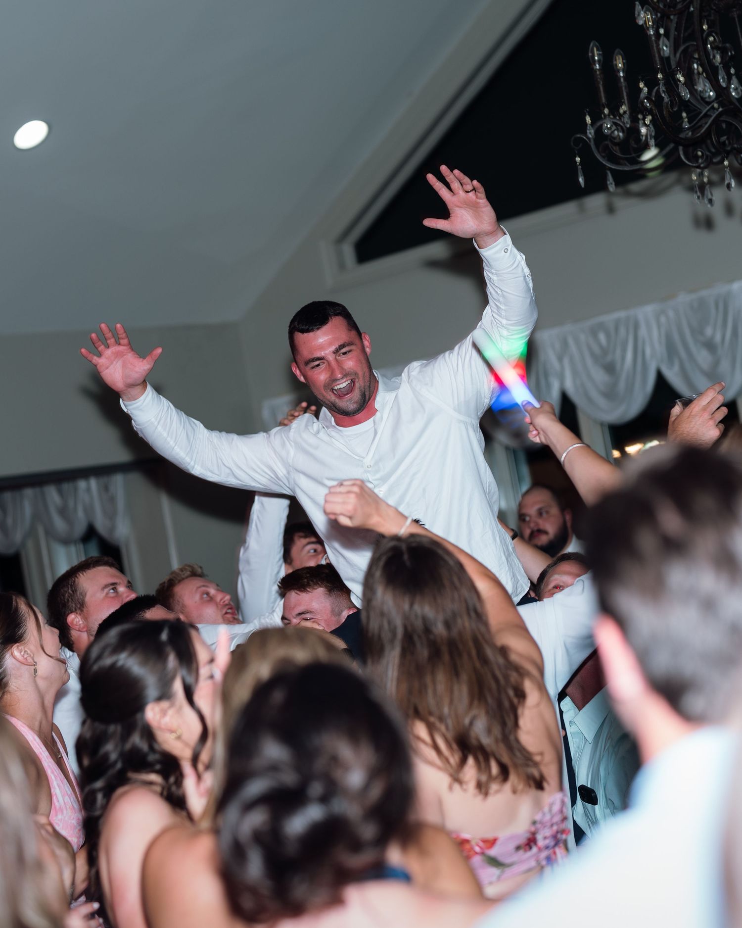 A groom in white celebrates on the dance floor, arms raised joyfully, surrounded by wedding guests at a lively reception.