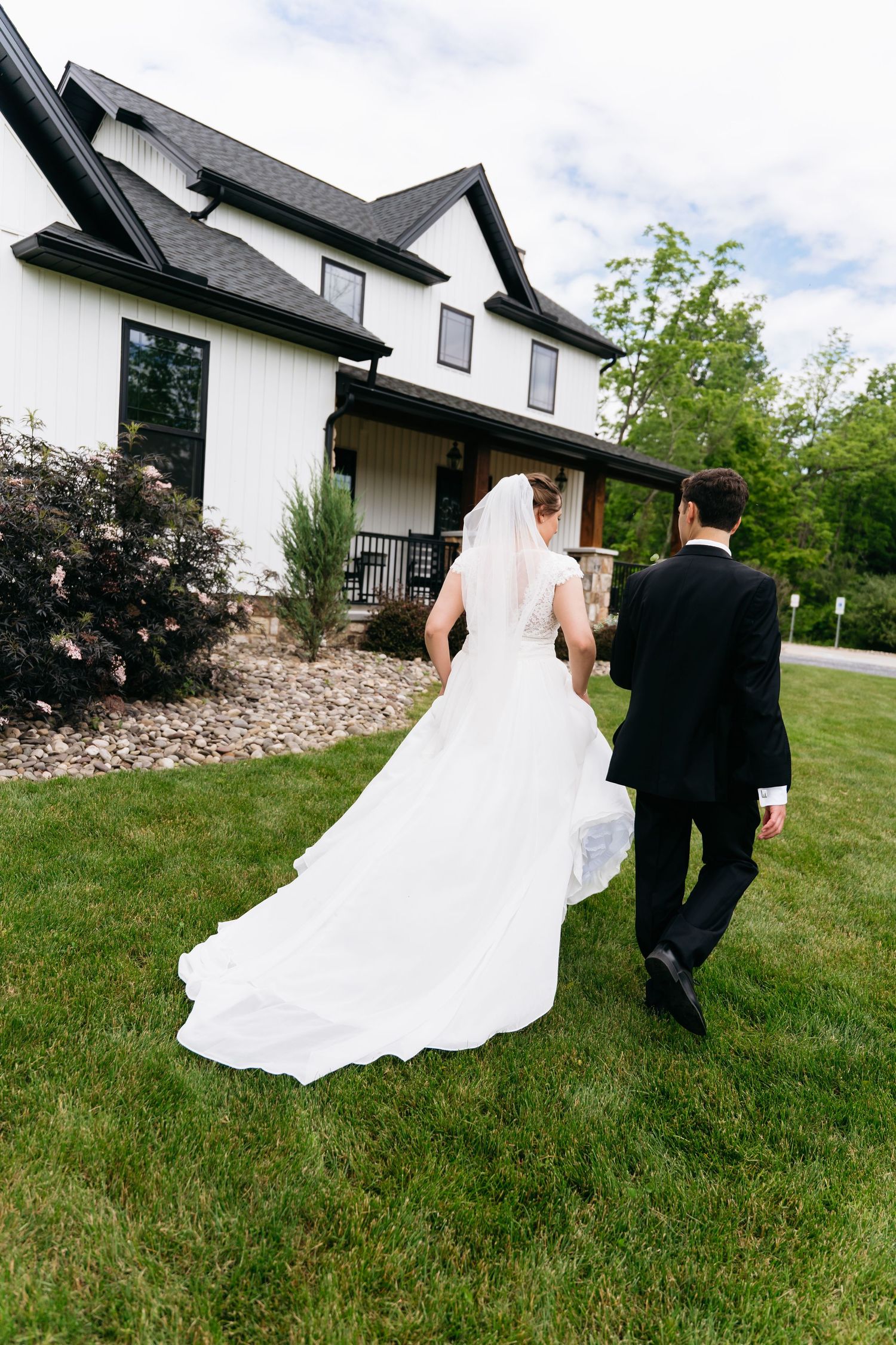 A couple in wedding attire walks across a grassy lawn with a modern white house in the distance.