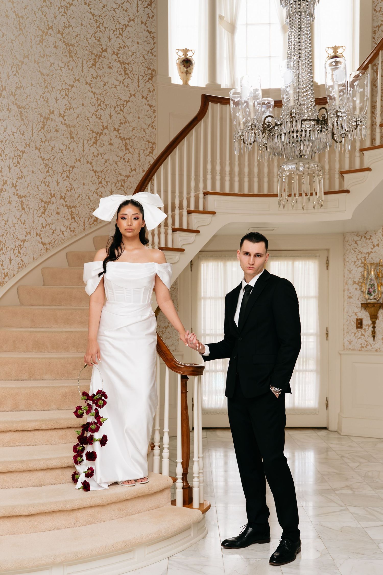 Bride in white gown and bow hat holds bouquet on staircase with groom in black suit in elegant mansion foyer.