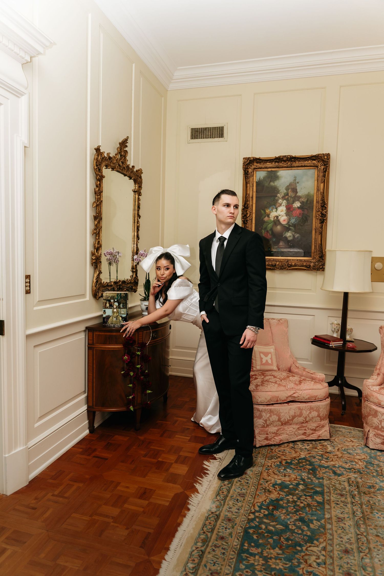 Elegant bride and groom posing in a classic room with ornate mirror, floral painting, and pink armchair.