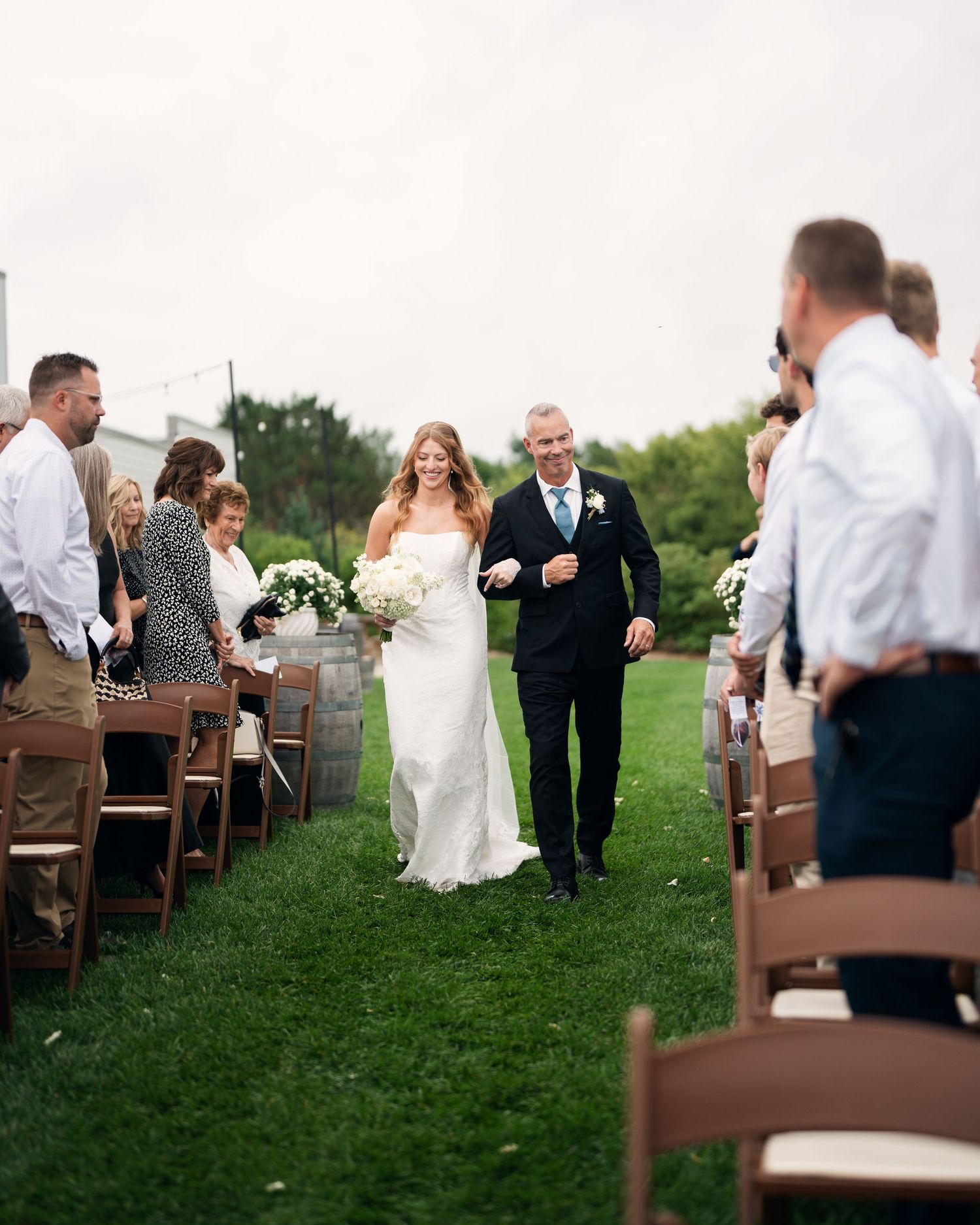 Smiling bride in white gown walks down outdoor aisle with escort past seated wedding guests on green lawn.