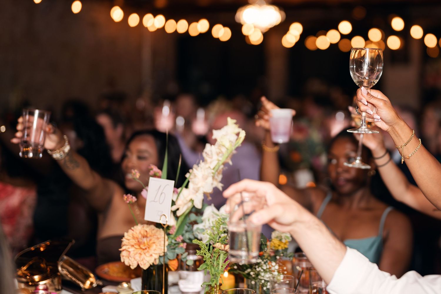 Guests raising wine glasses in a toast at an elegant wedding reception with floral centerpieces and warm string lights.