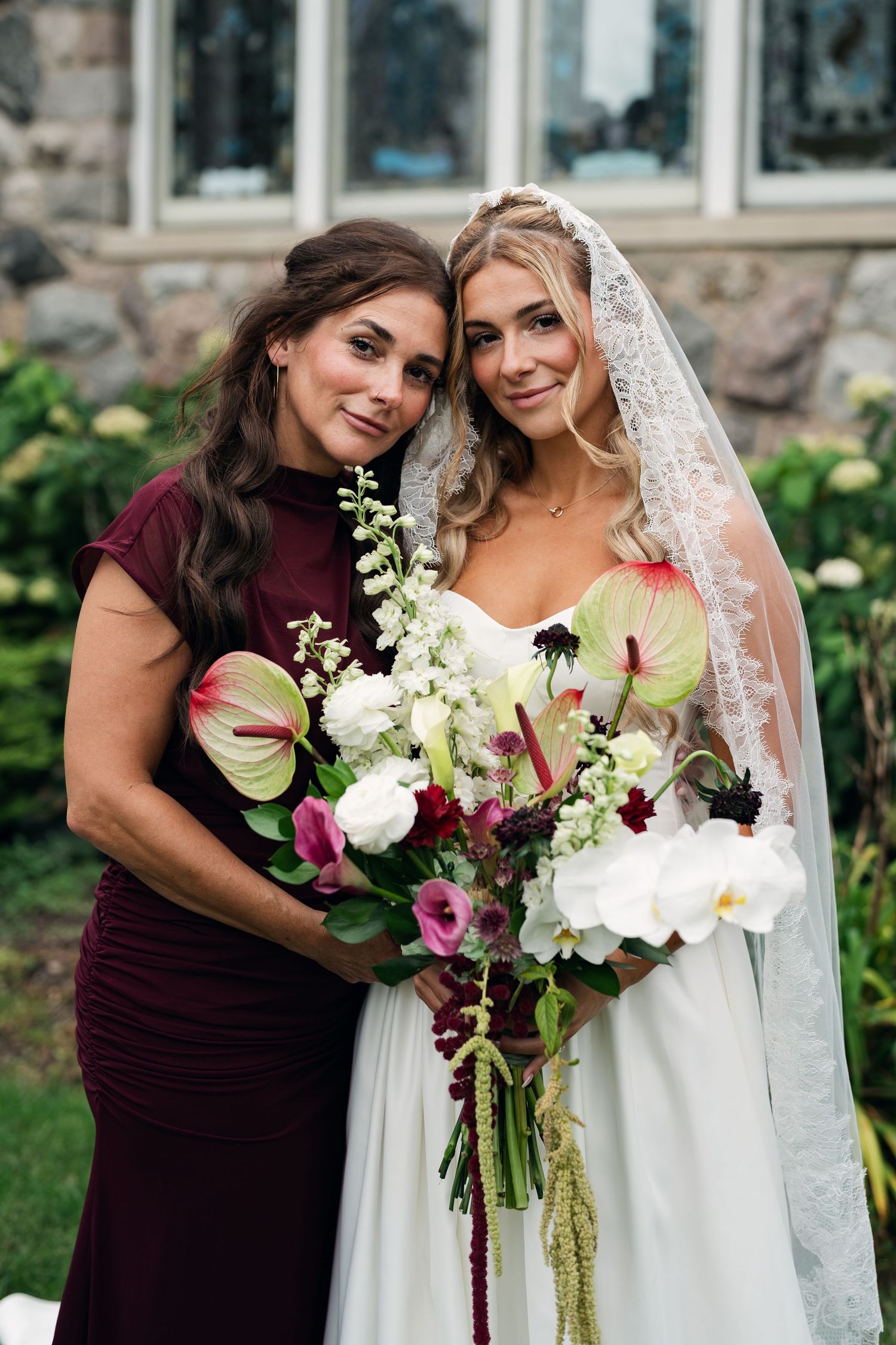 Bride in white gown and veil holds floral bouquet with bridesmaid in burgundy dress outdoors.