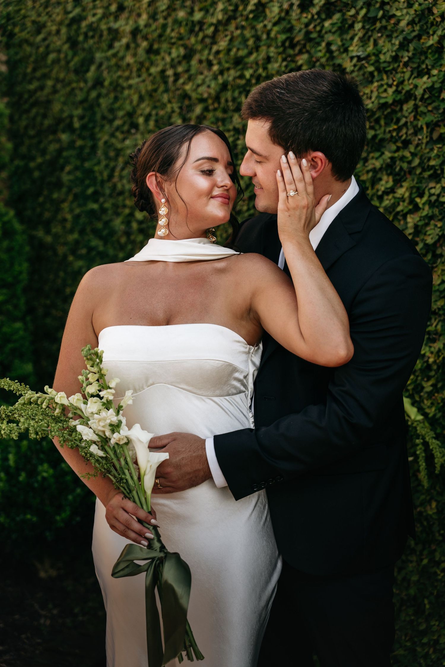A romantic wedding couple embraces against a lush green hedge backdrop during their outdoor photo session.