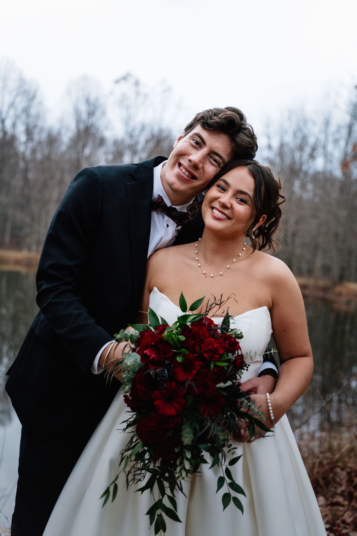 A couple shares joyful moments in their wedding attire with deep red rose bouquet against a winter landscape.