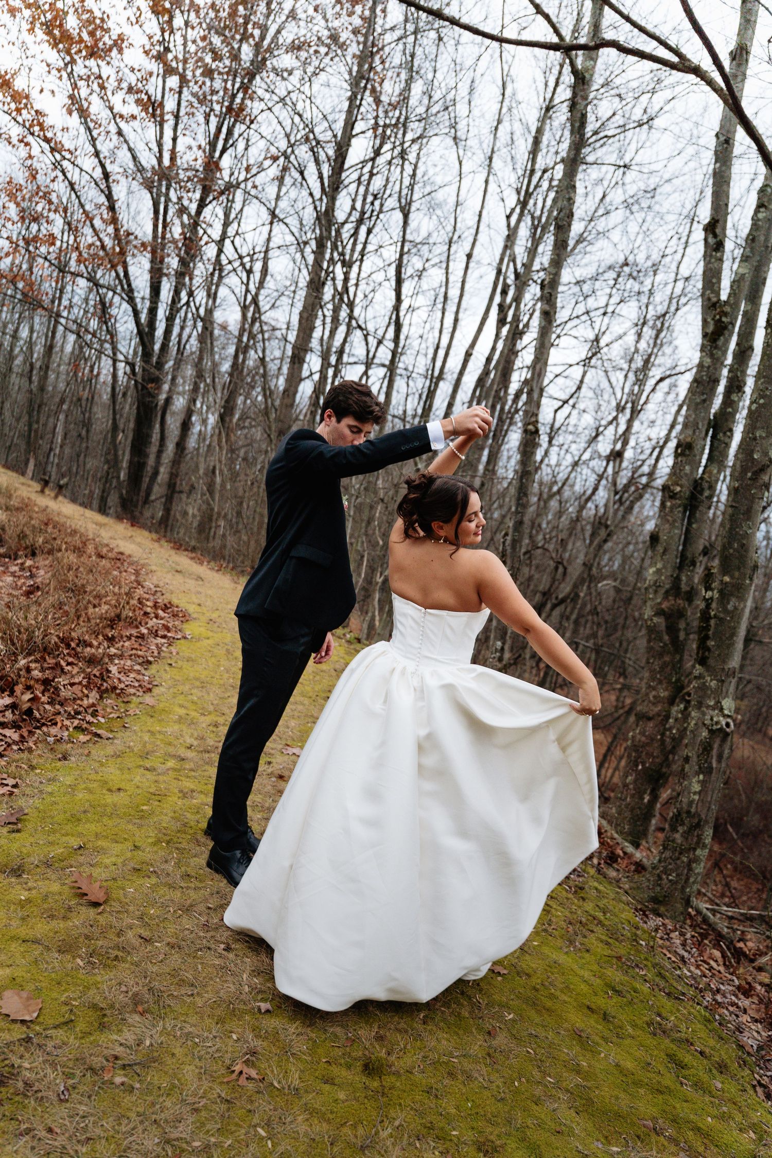 A couple in wedding attire dances together on a mossy path surrounded by bare autumn trees in the woods.