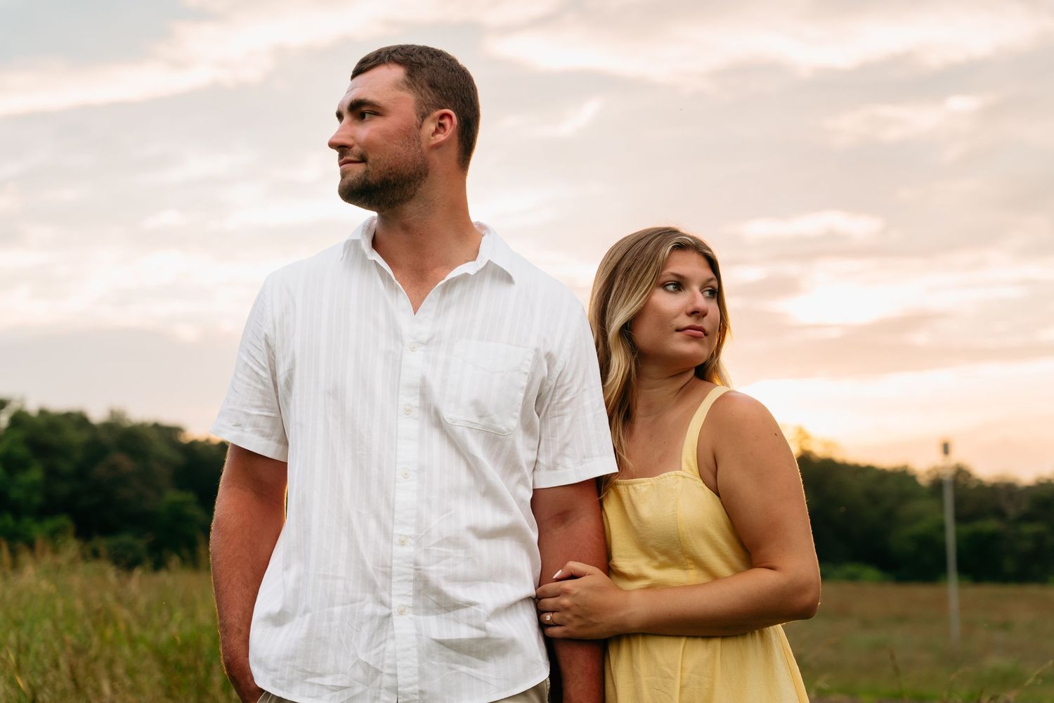 Couple in white and yellow outfits embrace while standing in a grassy field at sunset.