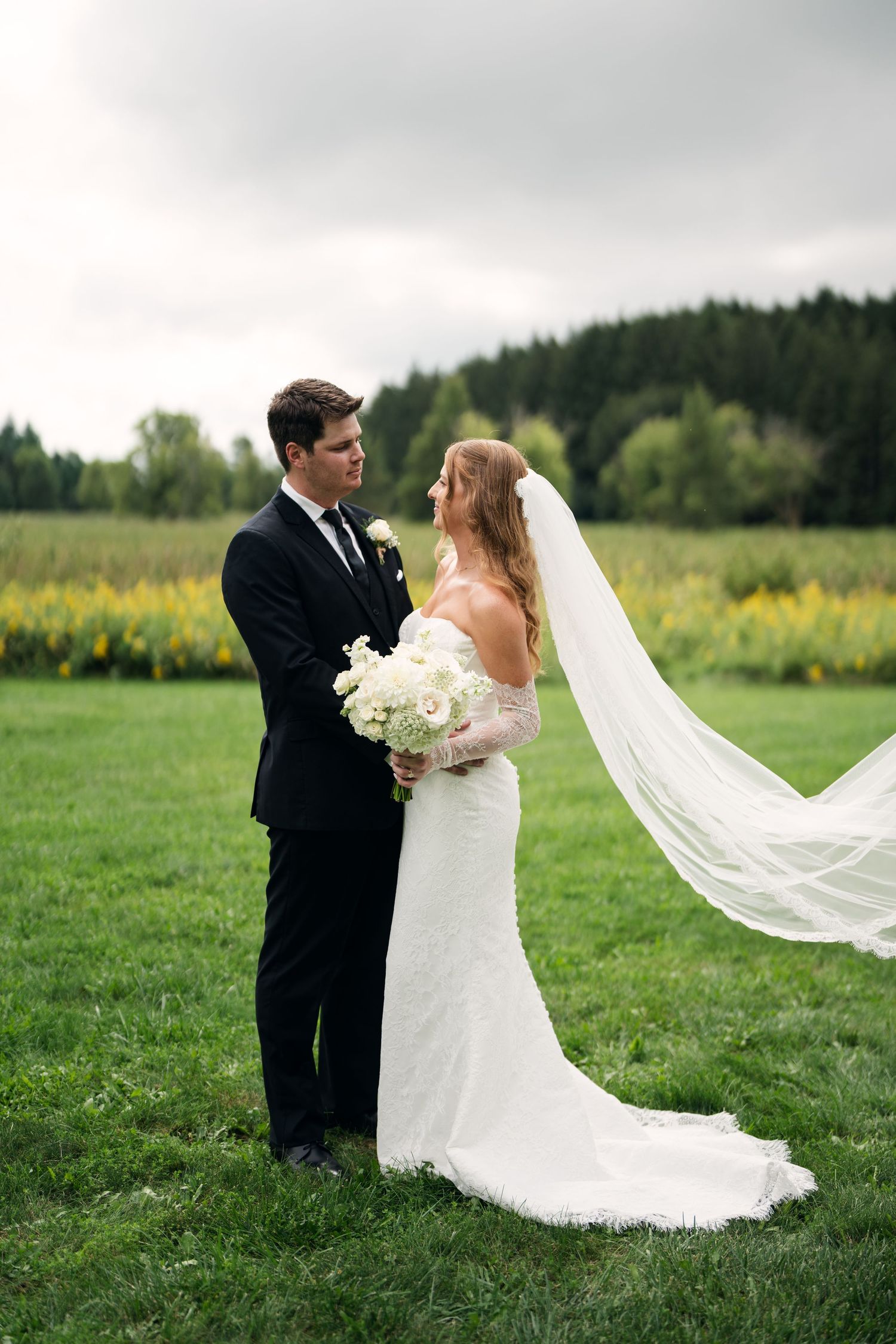 Bride in white lace gown with flowing veil holds bouquet while gazing at groom in black suit on green lawn.