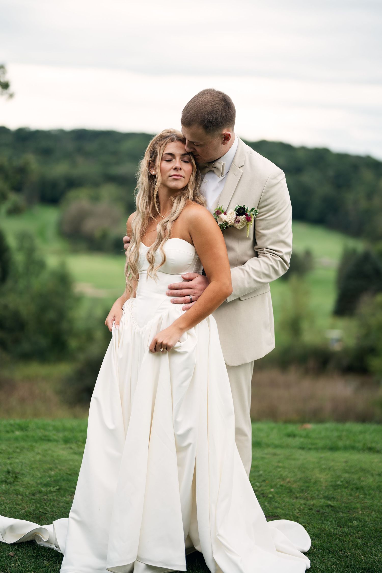 Bride in white gown and groom in beige suit embrace outdoors with lush green hills in the background.