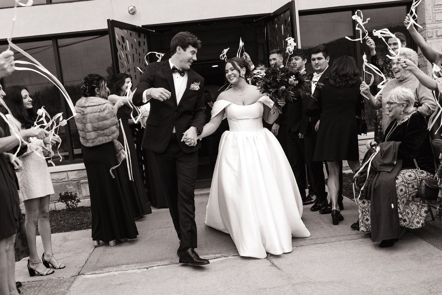 A black and white panoramic photo shows a wedding celebration with guests gathered outside a building.