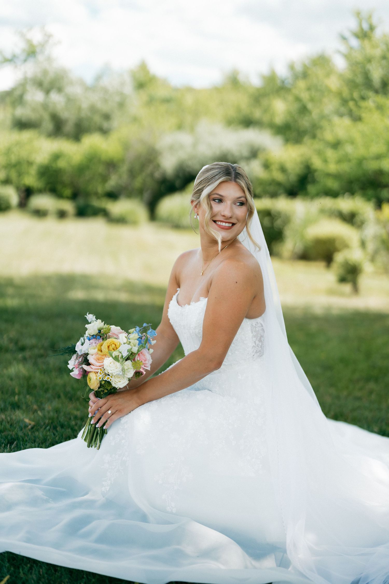 Smiling bride in white lace gown holding colorful bouquet while sitting on green grass outdoors.