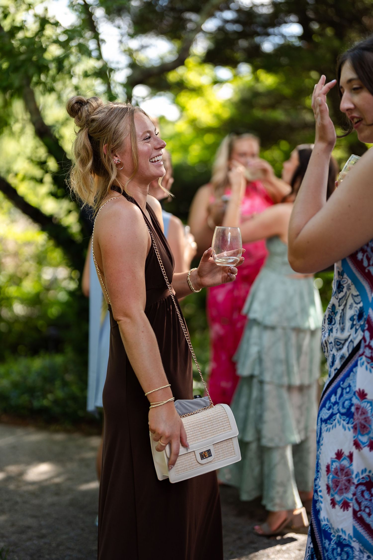 Laughing wedding guest in brown dress holding champagne and white purse at outdoor reception.