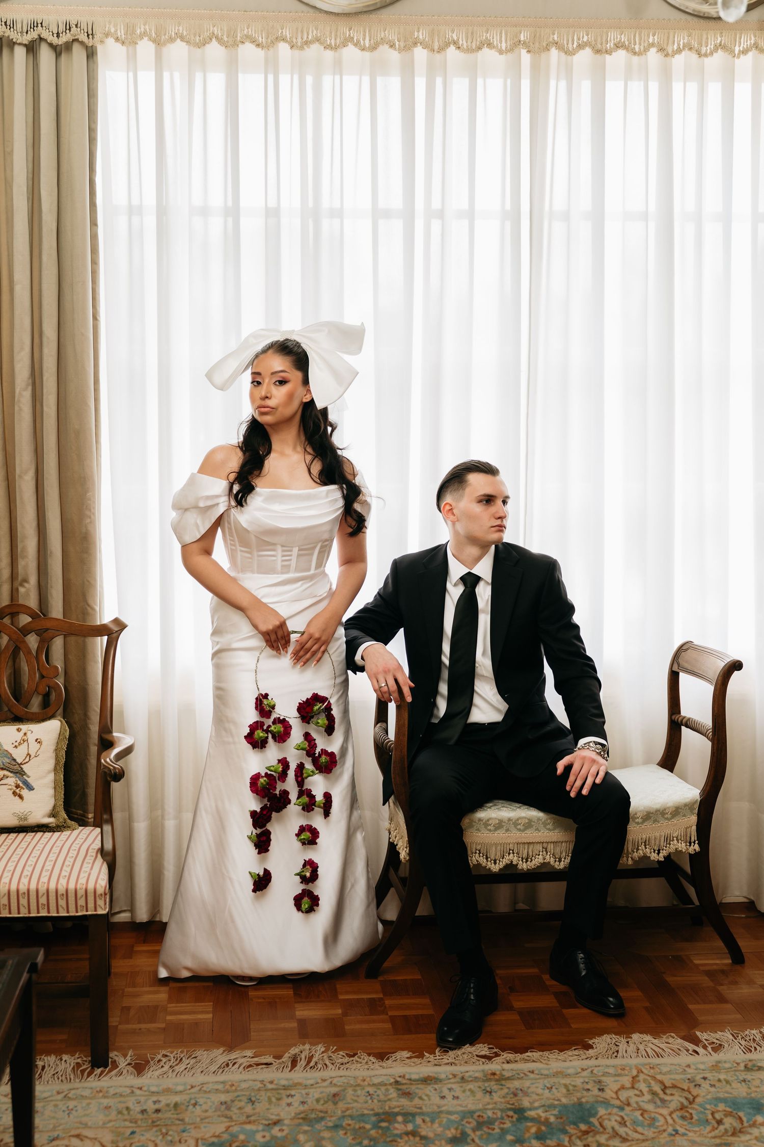 Bride in white gown with floral cascade and bow headpiece stands beside groom in black suit seated on vintage bench.