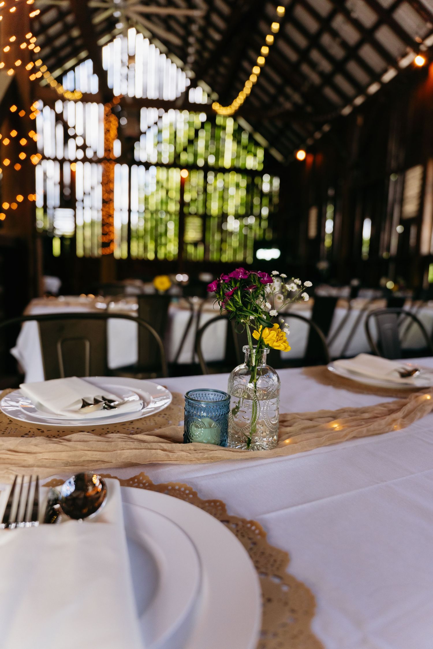 Rustic barn wedding reception table with vintage place settings, string lights, and wildflowers in glass vase.