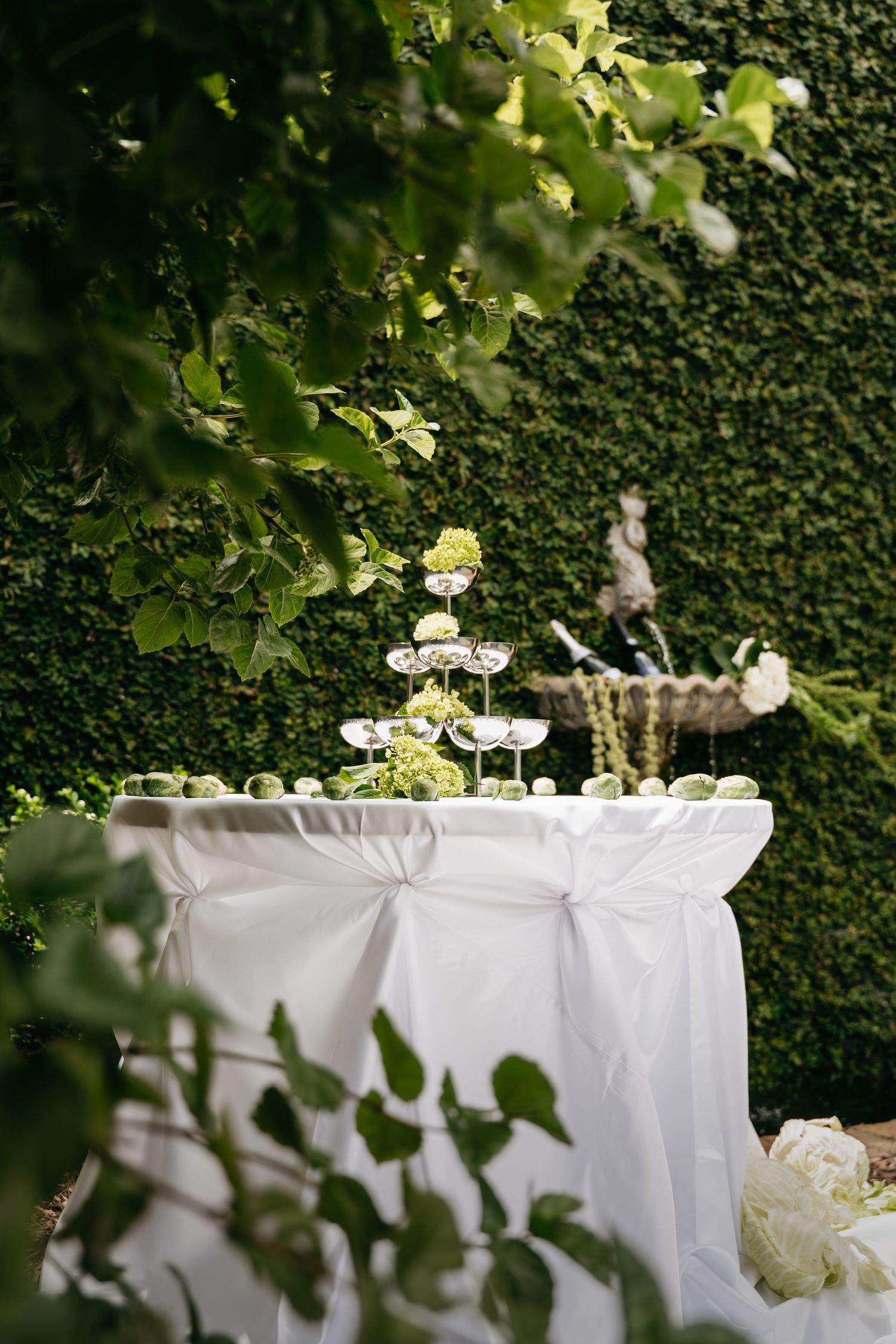 Elegant white tablecloth draped buffet table with floral arrangements set against a lush green hedge backdrop.