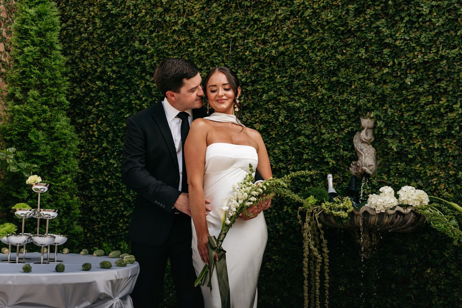 A wedding couple embraces in front of a lush green hedge wall during an elegant outdoor garden ceremony.
