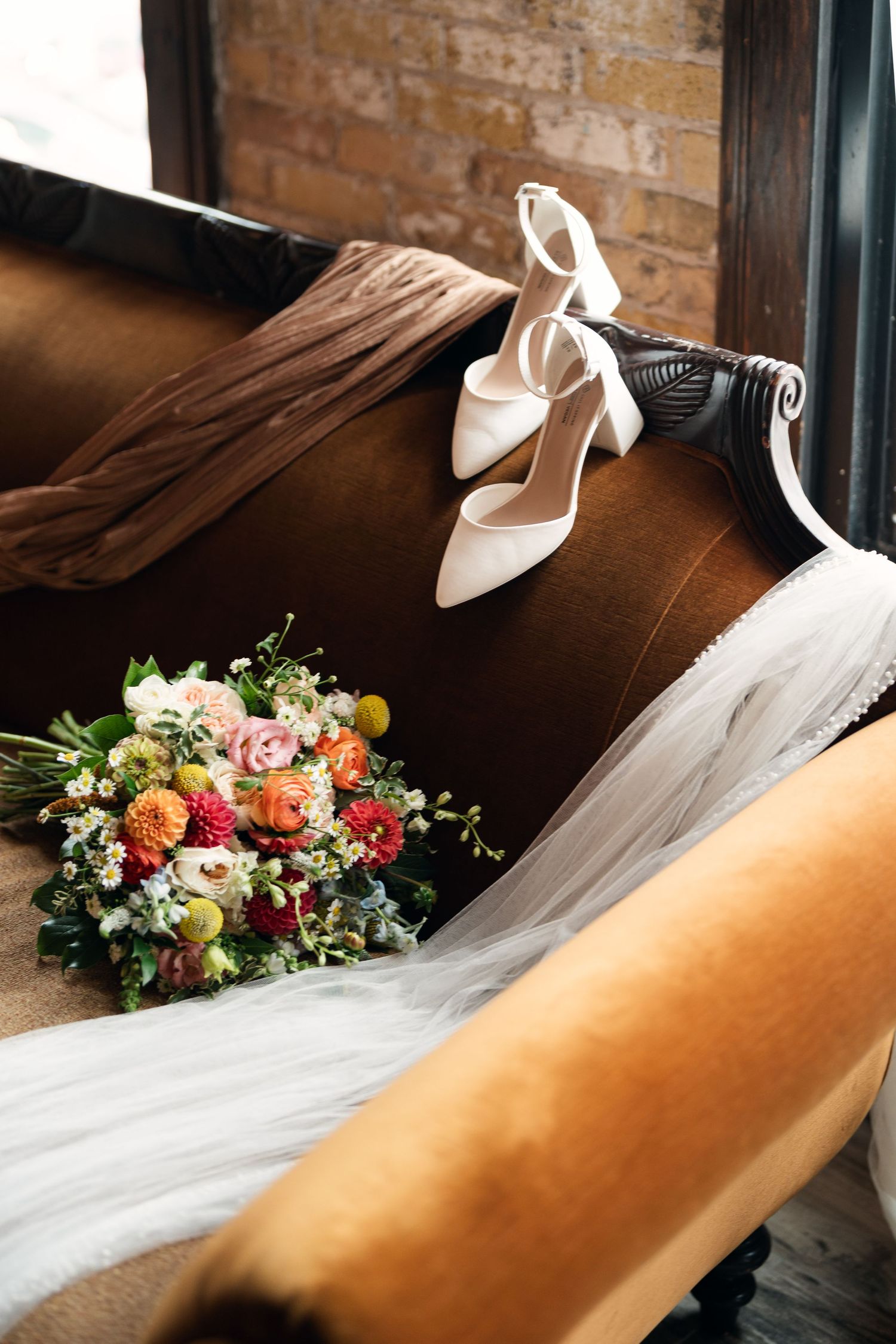 White strappy bridal heels and a vibrant mixed flower bouquet with veil on a brown leather vintage sofa.