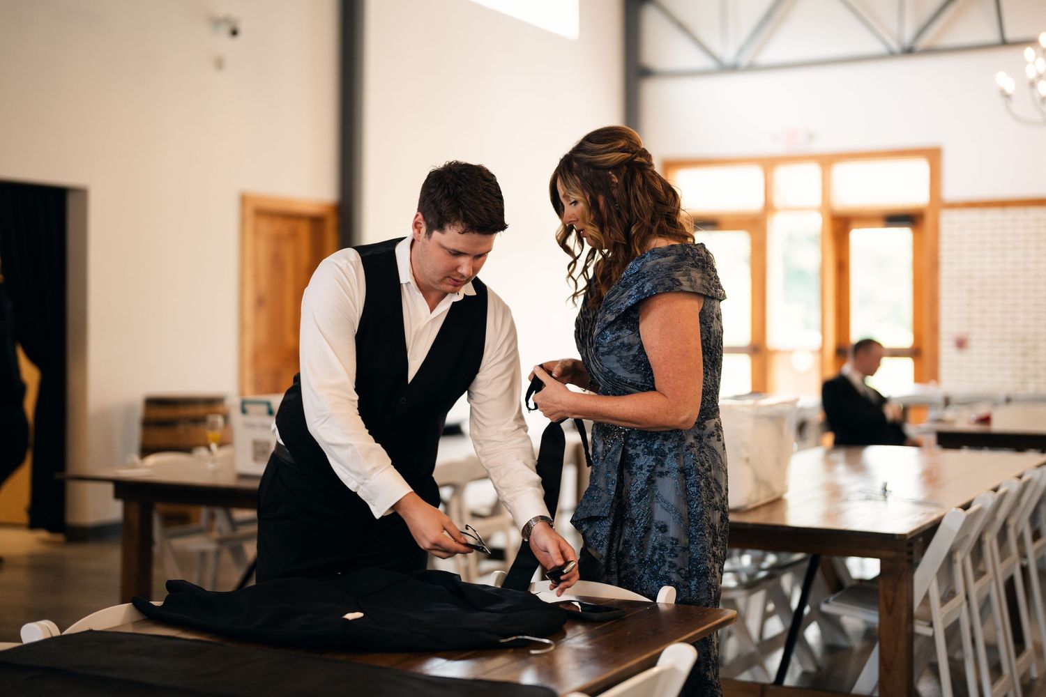 A man in a vest and woman in a blue dress arrange table linens at an elegant wedding venue.