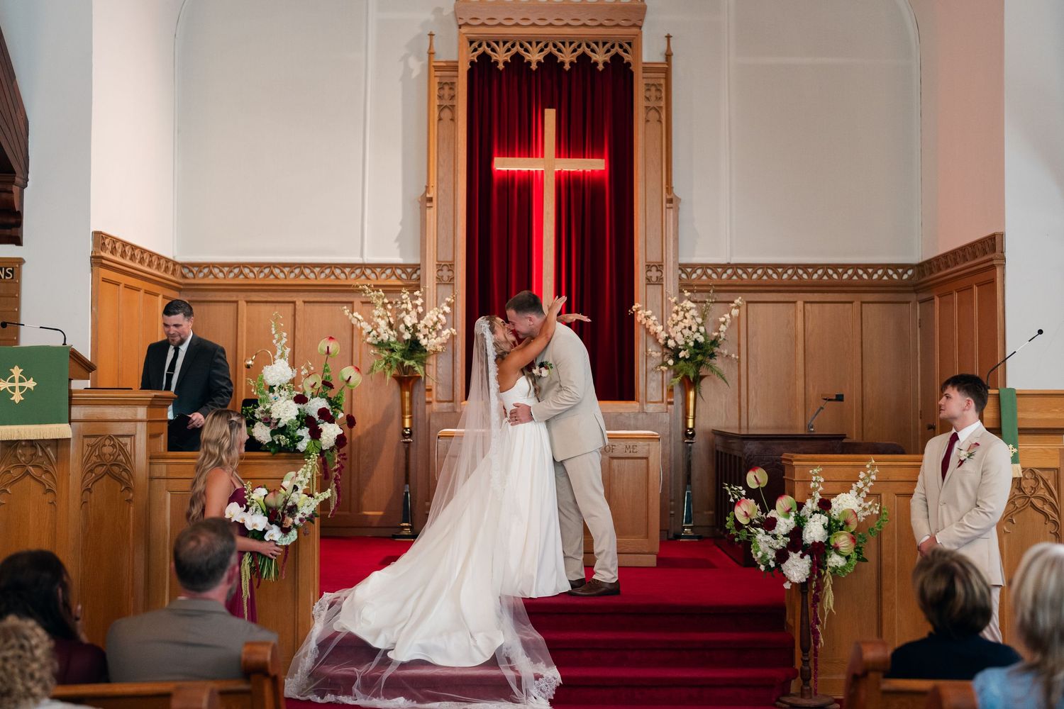 Bride and groom share first kiss at church altar adorned with floral arrangements and a glowing red cross.