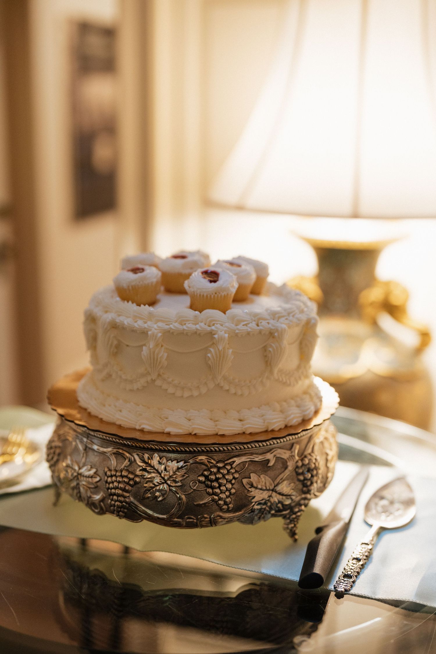 Two-tier white wedding cake with mini cupcakes on top displayed on ornate silver cake stand.