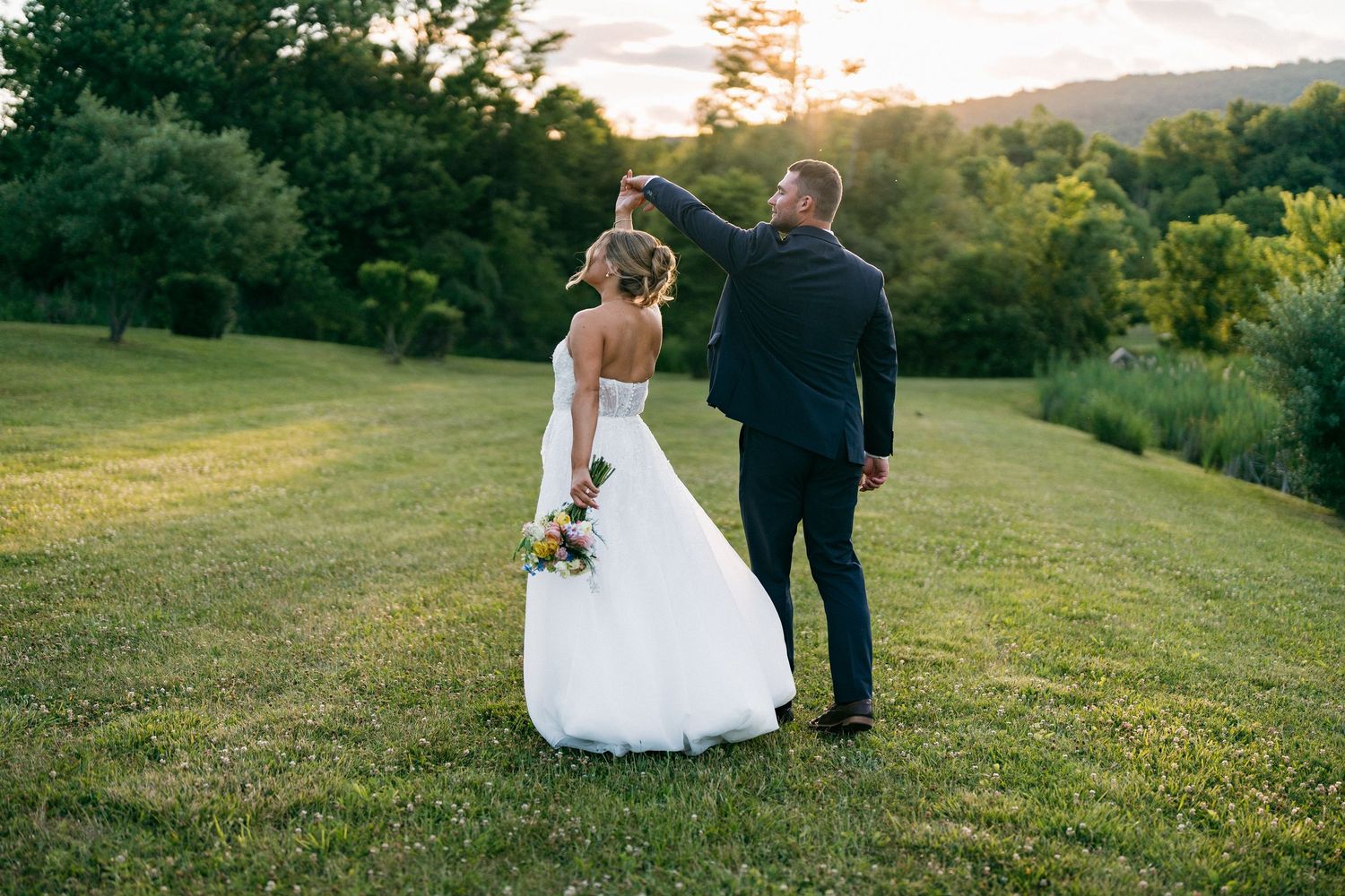Bride and groom dancing at sunset in a lush green field, with mountains and trees in the background.