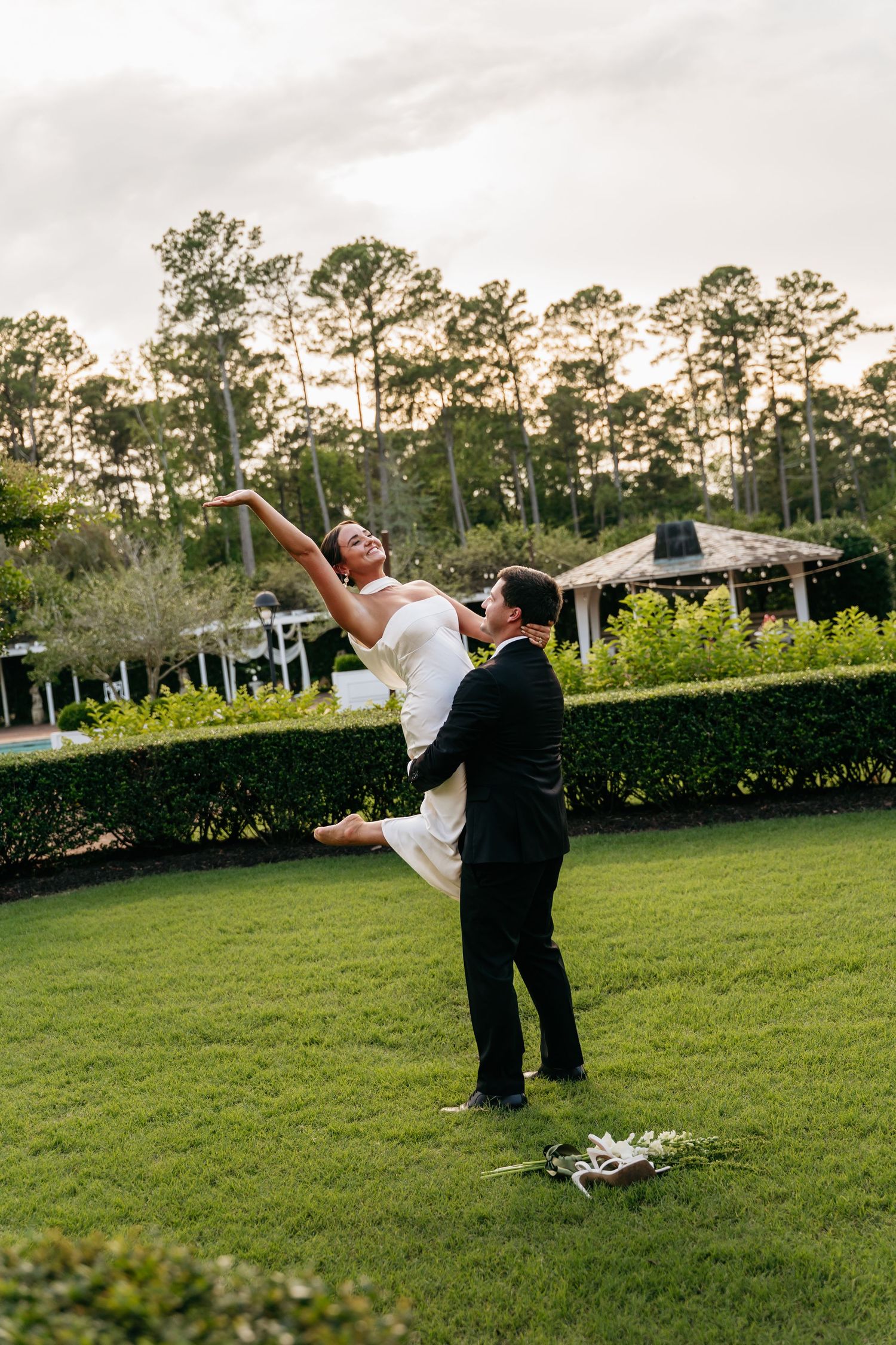A couple performs a dramatic dance lift pose on a lush green lawn at sunset with pine trees in the background.