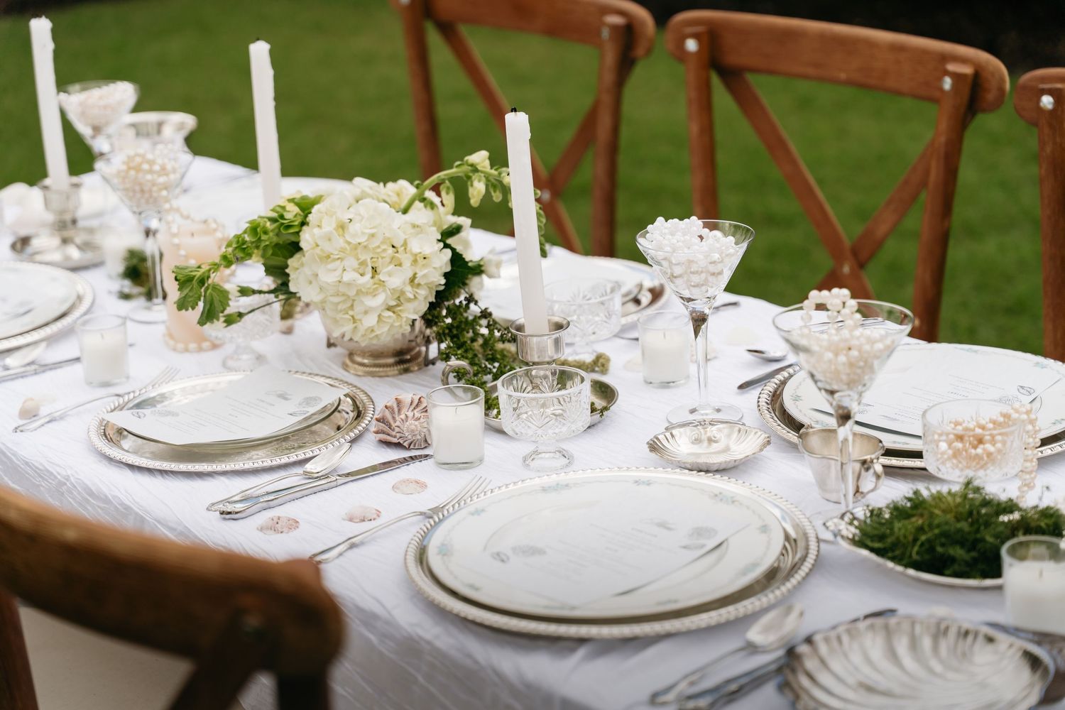 Elegant white table settings with floral centerpieces and crystal glassware arranged outdoors for a garden wedding reception.