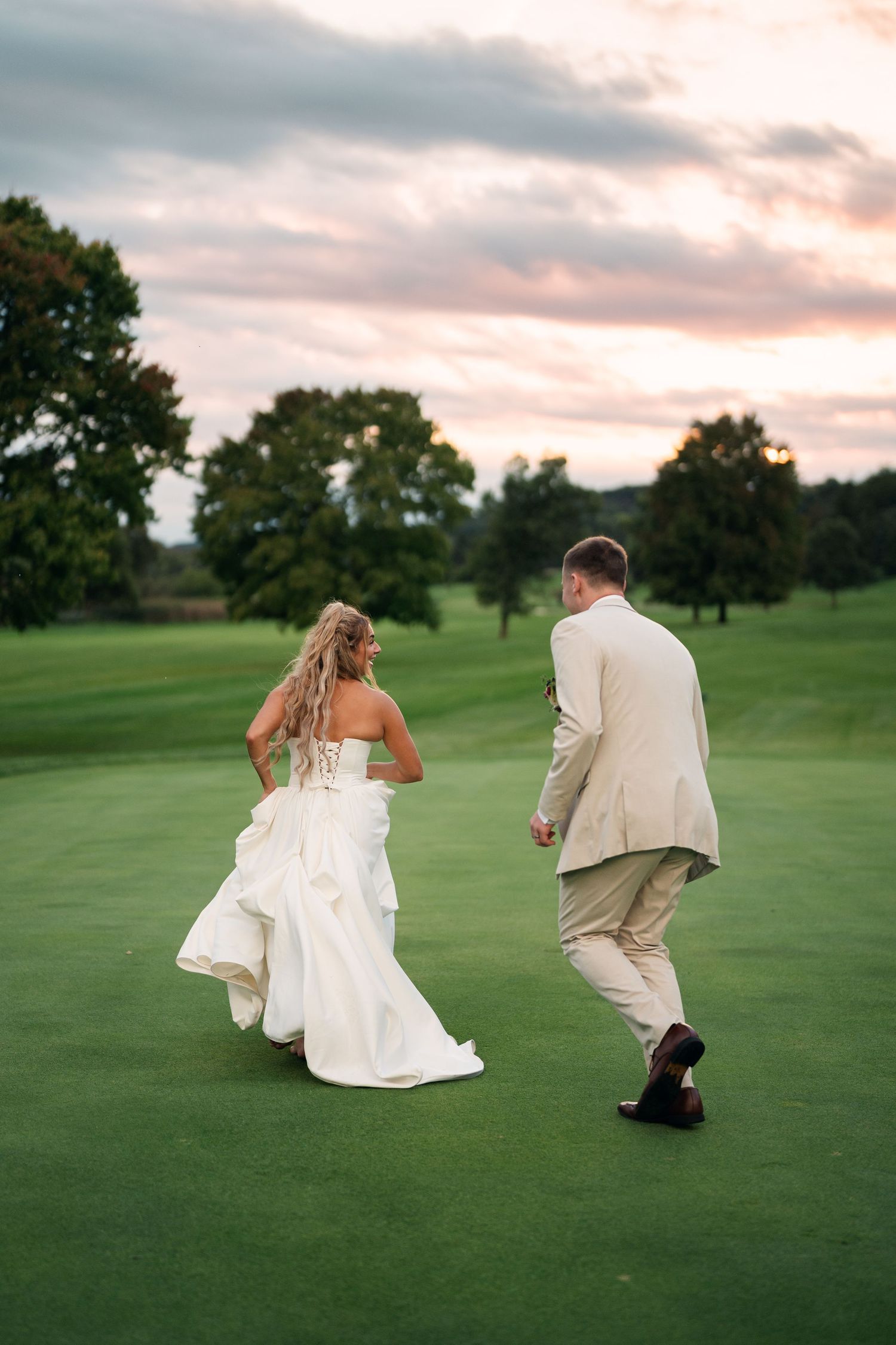 Bride and groom walk hand in hand across a lush green golf course at sunset in elegant wedding attire.