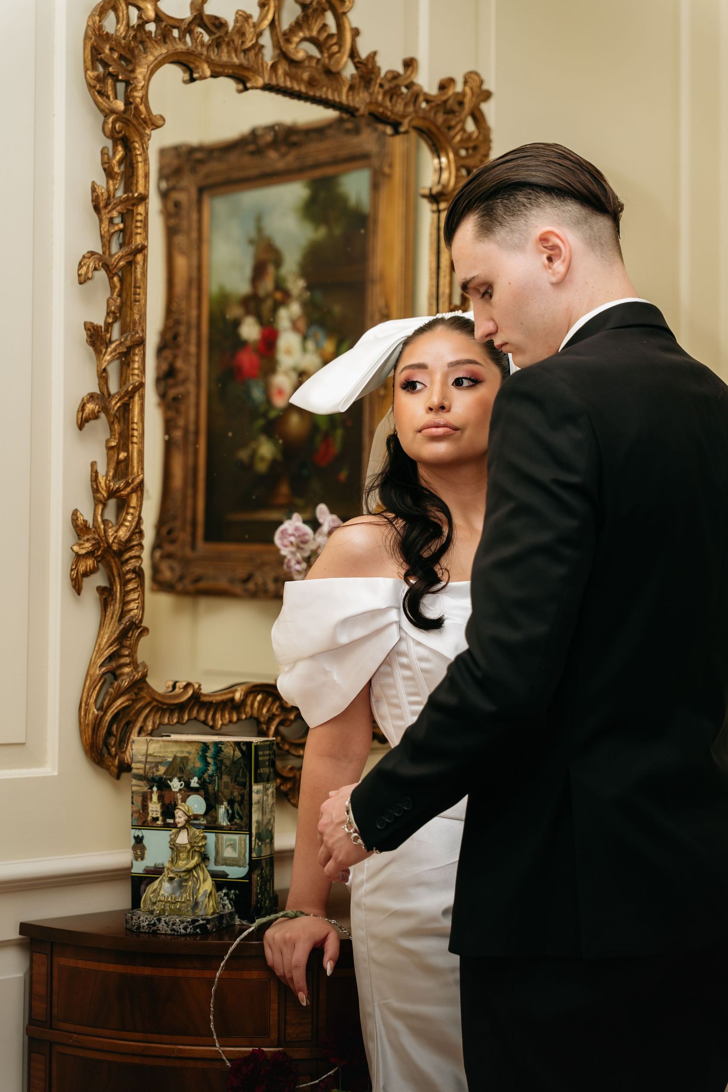 Bride in white off-shoulder dress with bow hat and groom in black suit stand before ornate gold-framed painting.