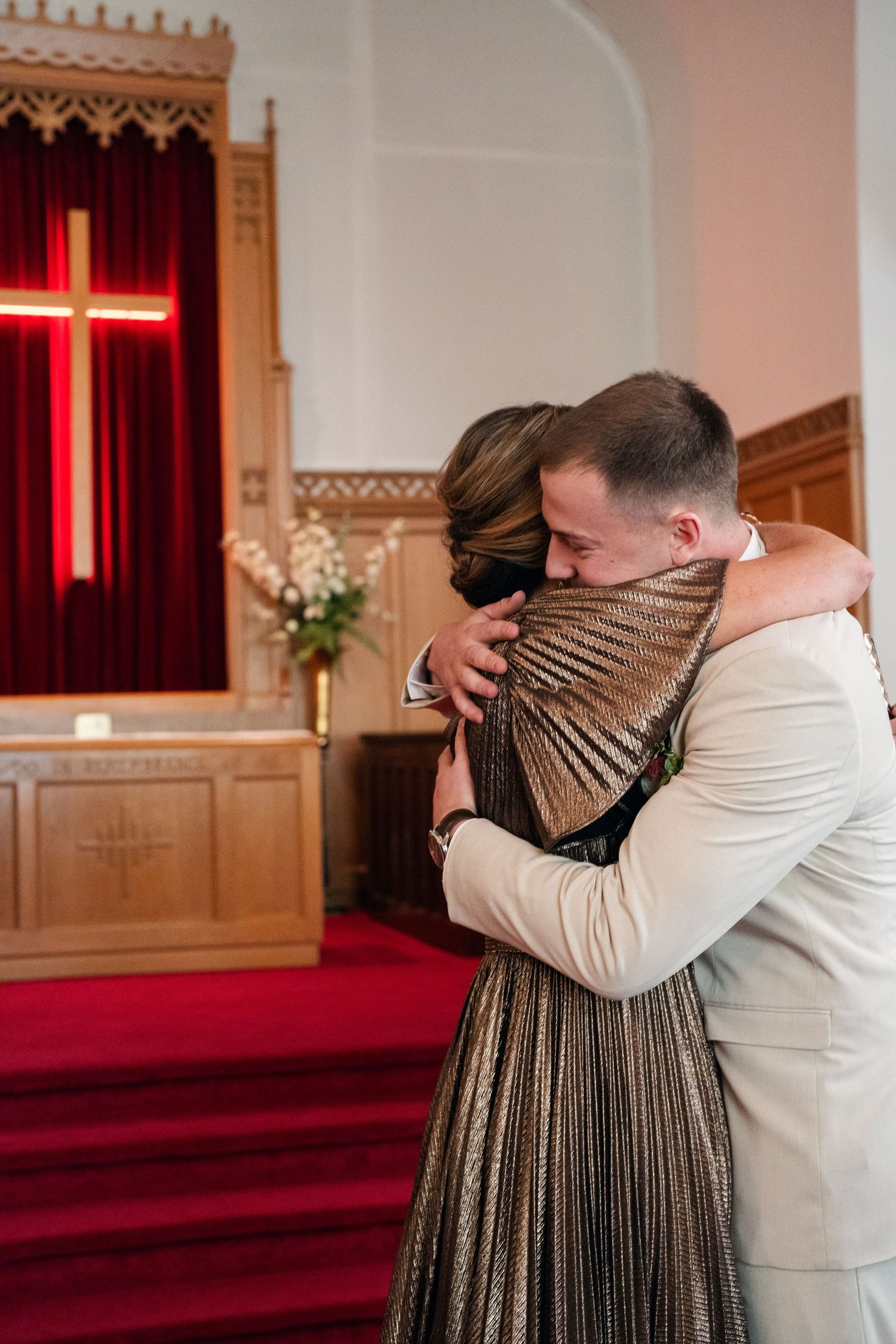 Groom in beige suit embracing a woman in a gold dress inside a church with a glowing cross altar.