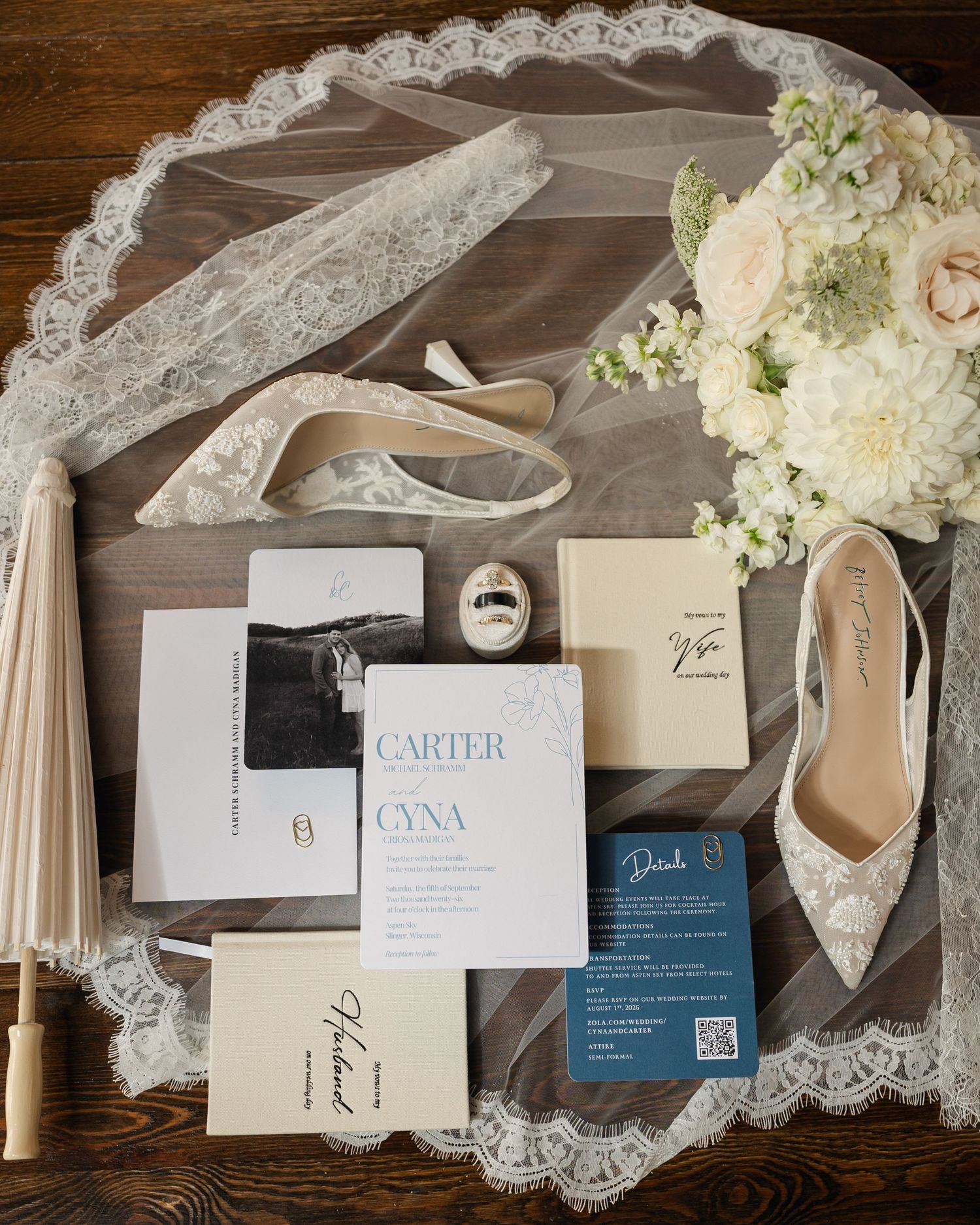 Elegant wedding flat lay featuring lace veil, bridal heels, invitation suite, and white floral bouquet on wood background.