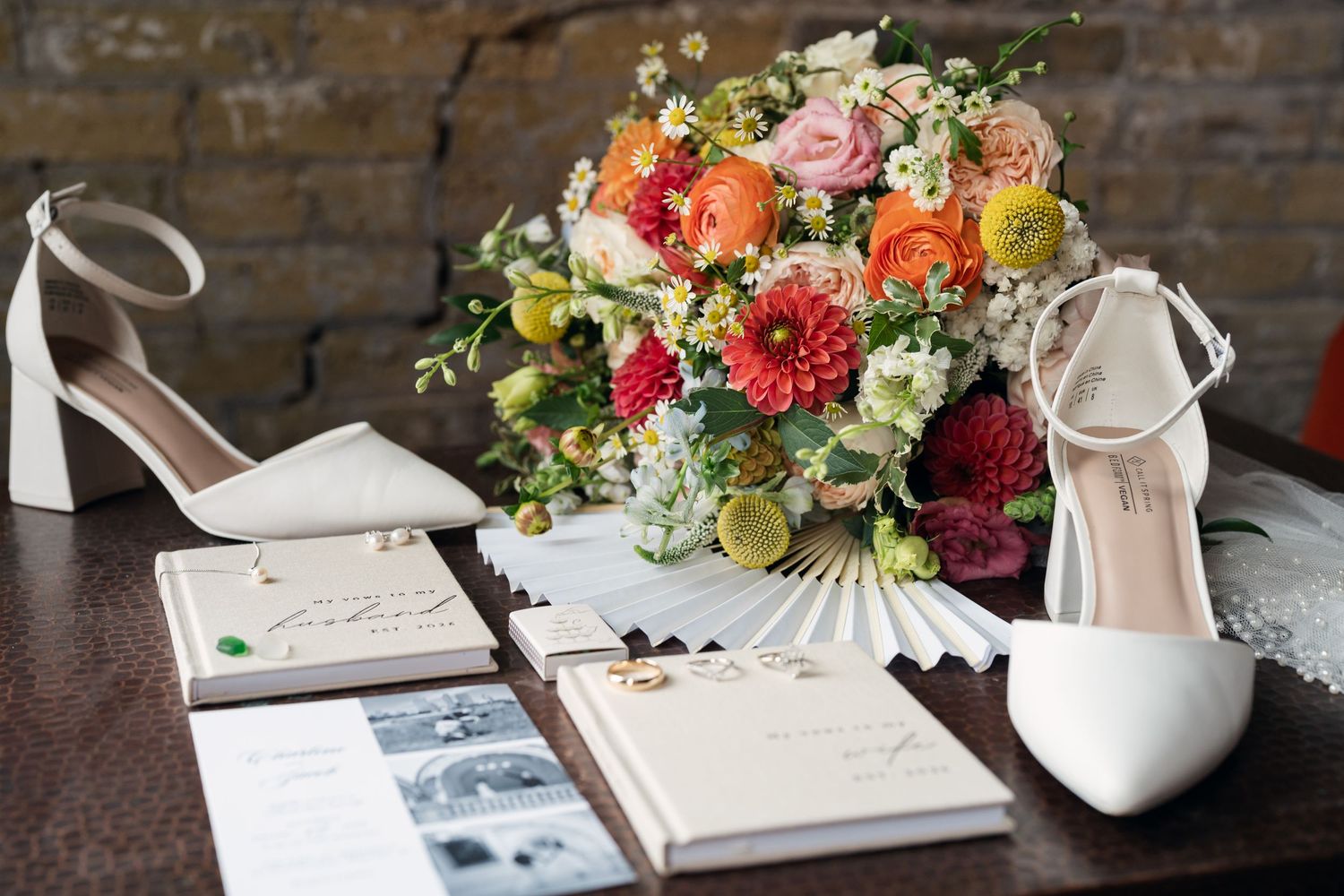 White bridal heels, colorful wedding bouquet with dahlias and ranunculus, and wedding stationery arranged on a wooden table.