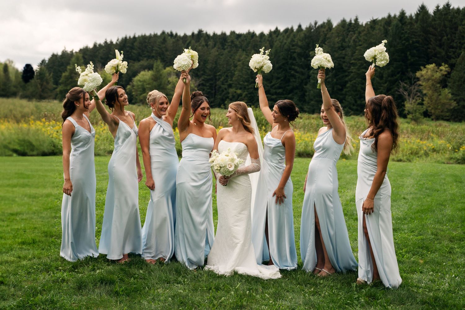 Bride with six bridesmaids in light blue gowns raising white bouquets on a lush green lawn.