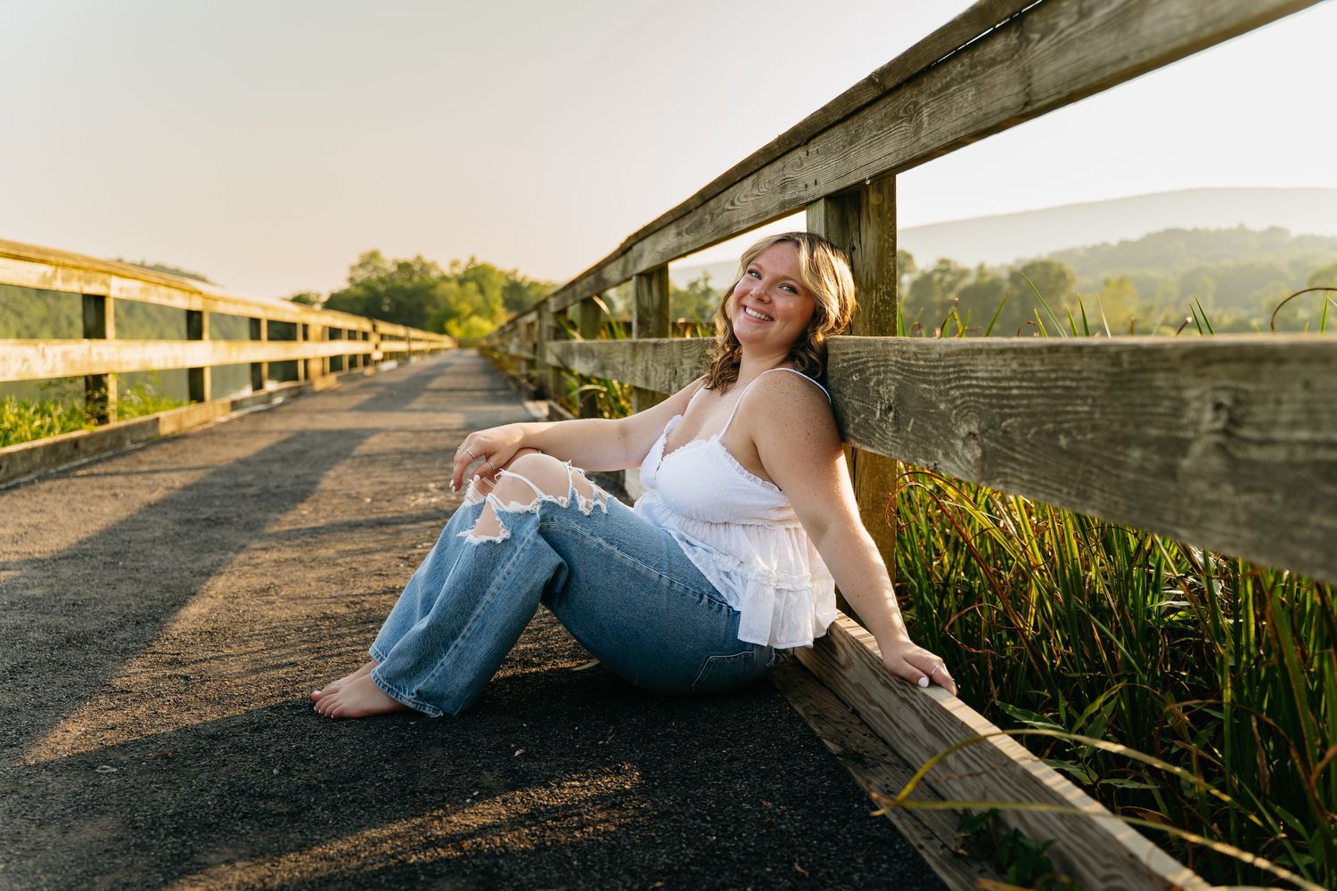 Young woman sitting on a wooden bridge path at golden hour, smiling in white top and jeans.