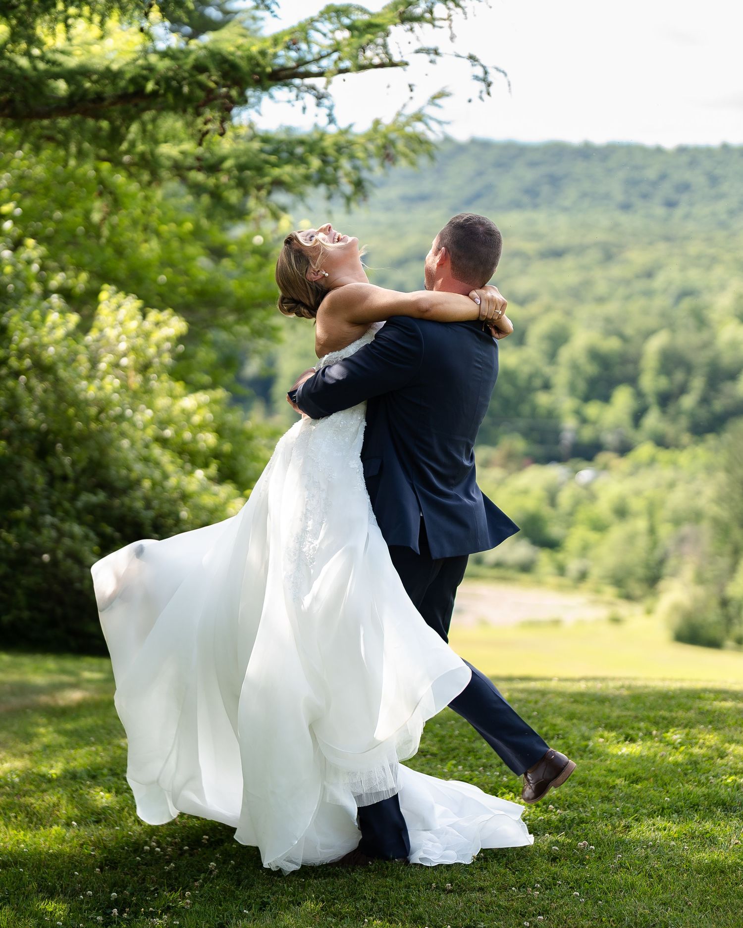 Groom lifts bride in white wedding gown outdoors, both laughing joyfully with green hills behind them.