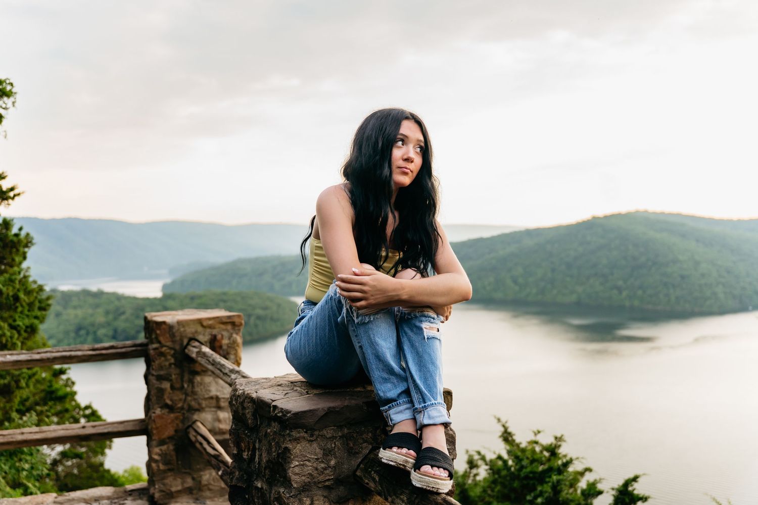 Young woman with dark hair sits on stone wall overlooking scenic river valley with green mountains and cloudy sky.