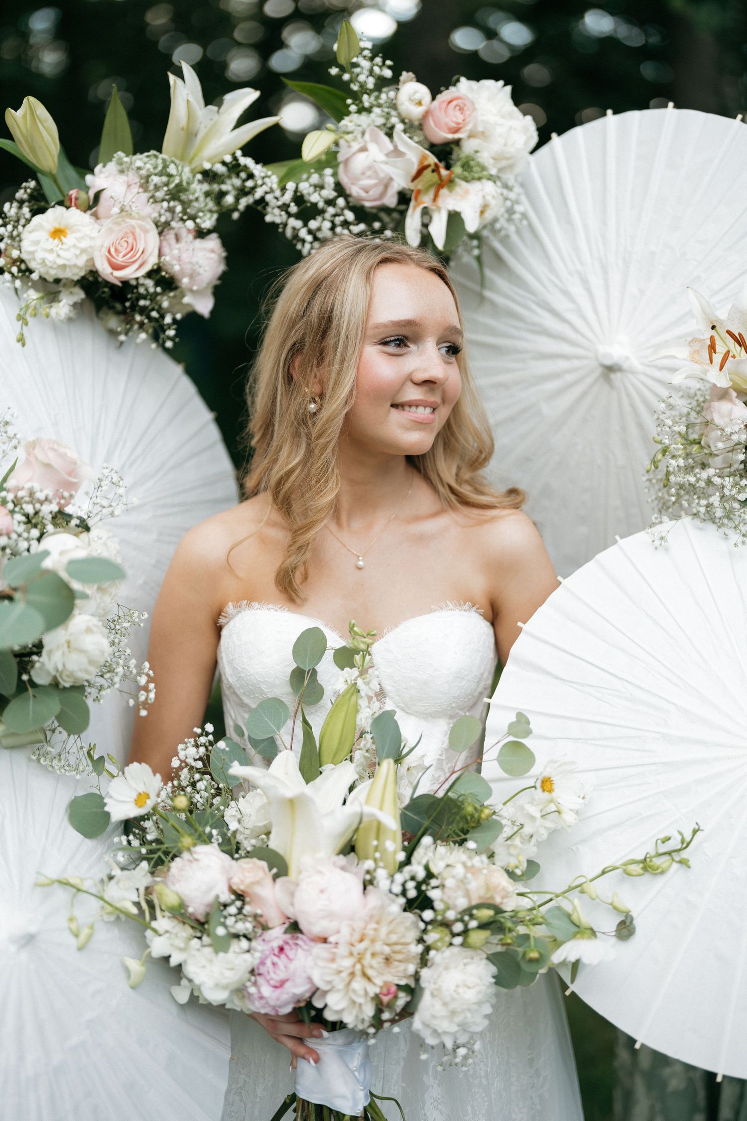 Young bride in strapless white gown holds lush floral bouquet before paper fan backdrop with pink and white flowers.