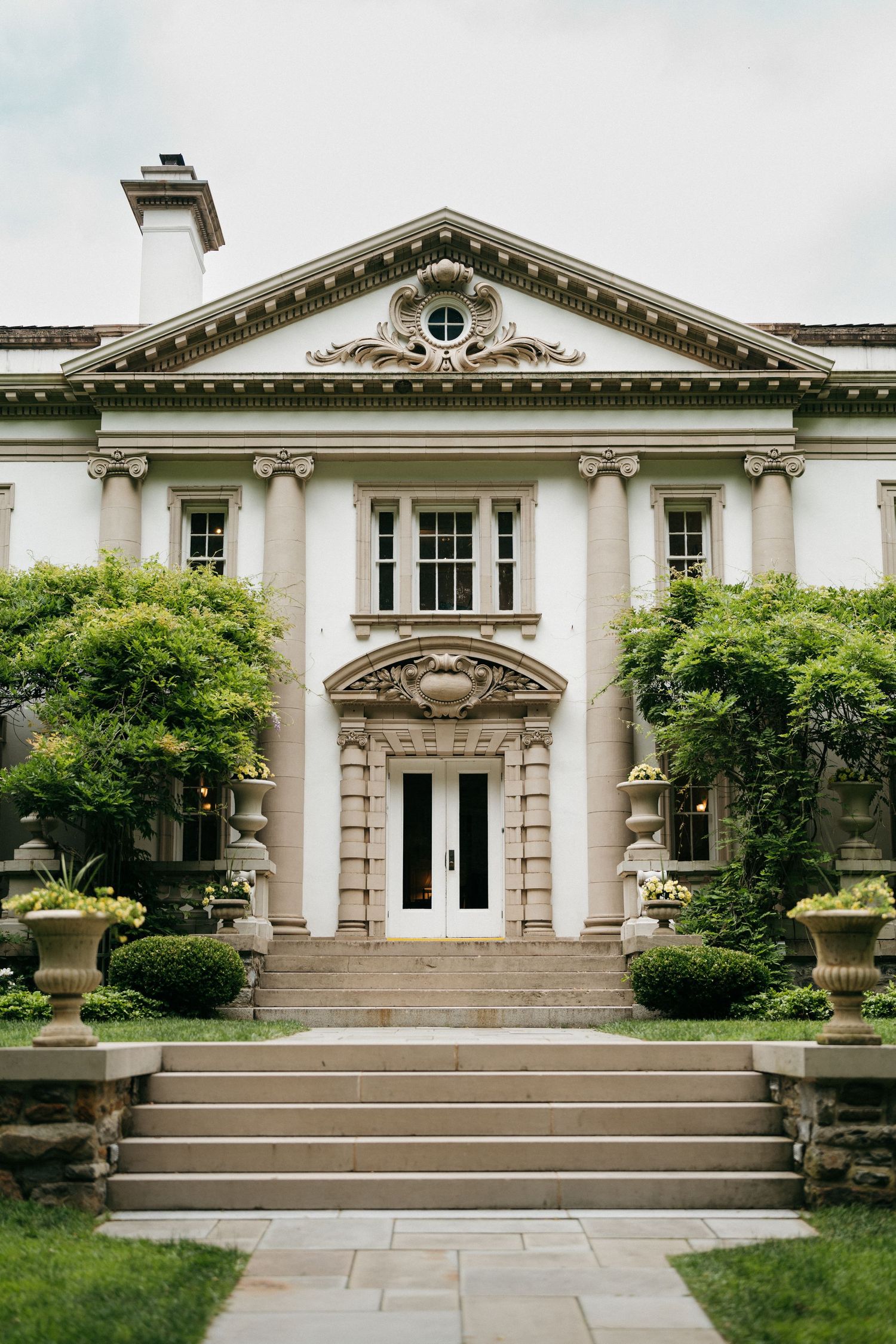 Elegant neoclassical mansion with white columns, ornate pediment, grand entrance steps, and lush green landscaping.
