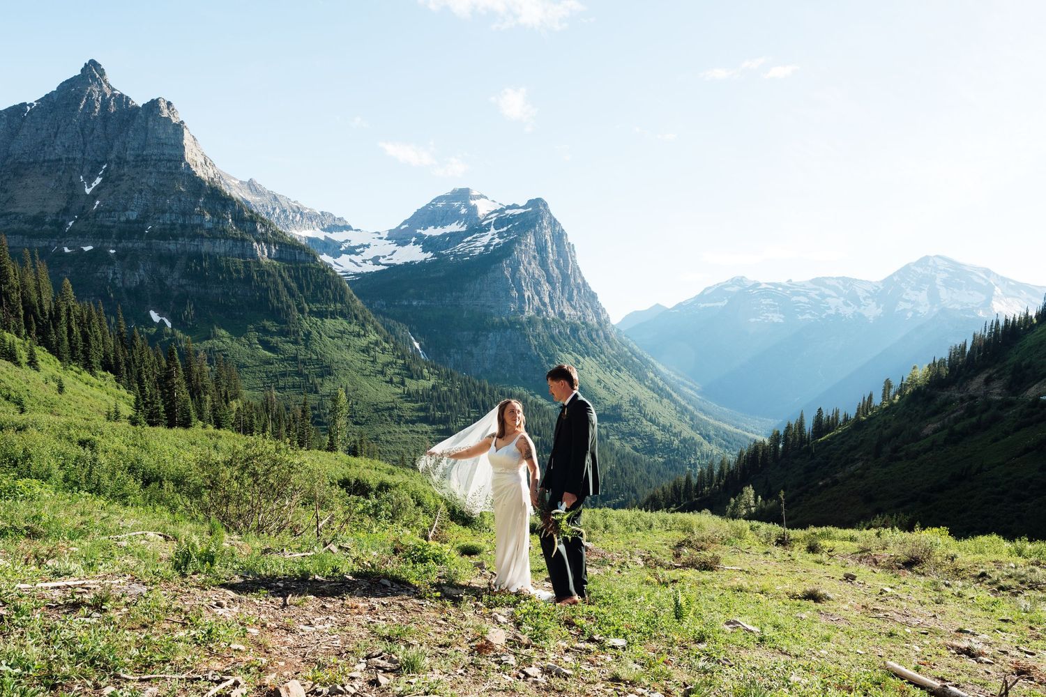 Bride and groom stand on a scenic mountain meadow with stunning peaks and glaciers in Glacier National Park.